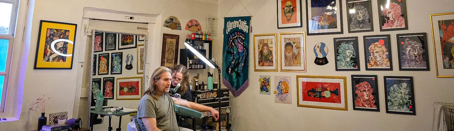 A tattoo artist tattooing a man's arm inside an art studio with colorful framed artwork on the walls.