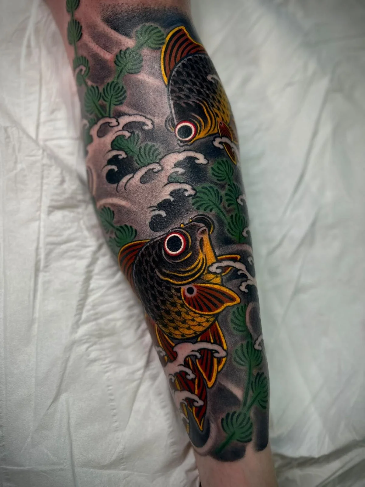 Tattoo of two koi fish surrounded by green leaves and waves on a person's arm.