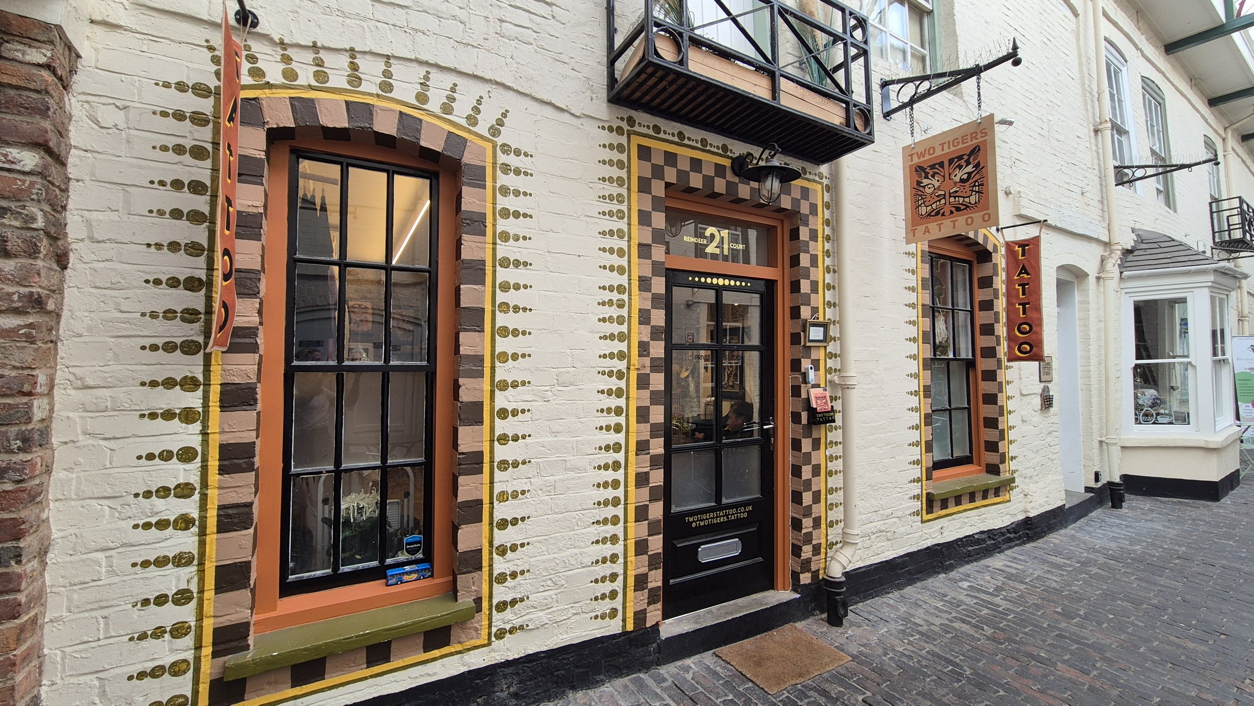 The exterior of a tattoo shop with a textured white brick wall, decorated with black and brown checkered and dotted designs around the windows and door. There are two windows with black frames, and a door with a black frame. Signage includes a vertical banner reading 'TATTOO', a hanging sign with a tiger's face labeled 'TWO TIGERS TATTOO,' and a website URL for the tattoo shop. The street is paved with cobblestones.