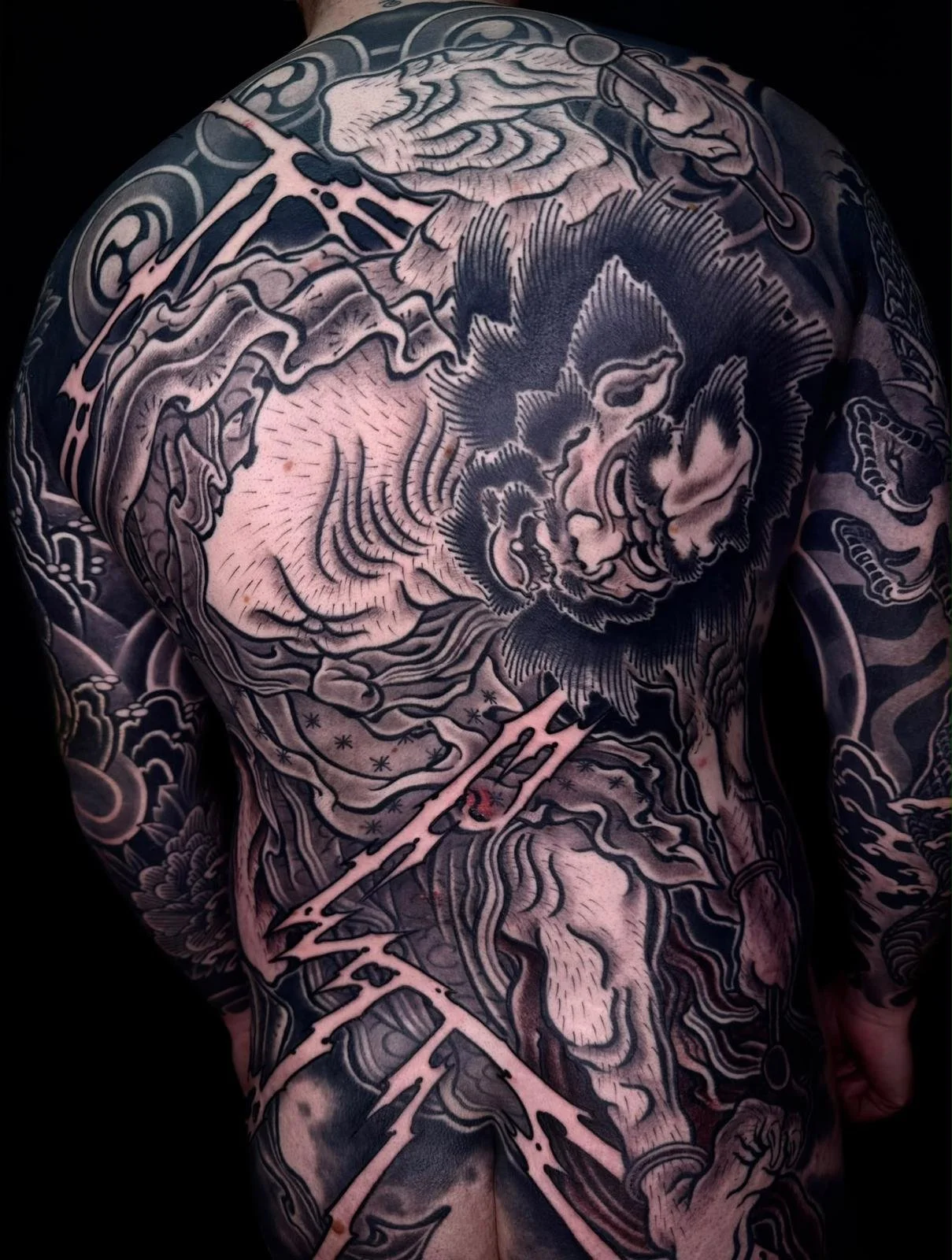 A person's back and arm covered in a detailed black and gray Japanese-style tattoo, featuring a fierce lion, clouds, and traditional patterns.