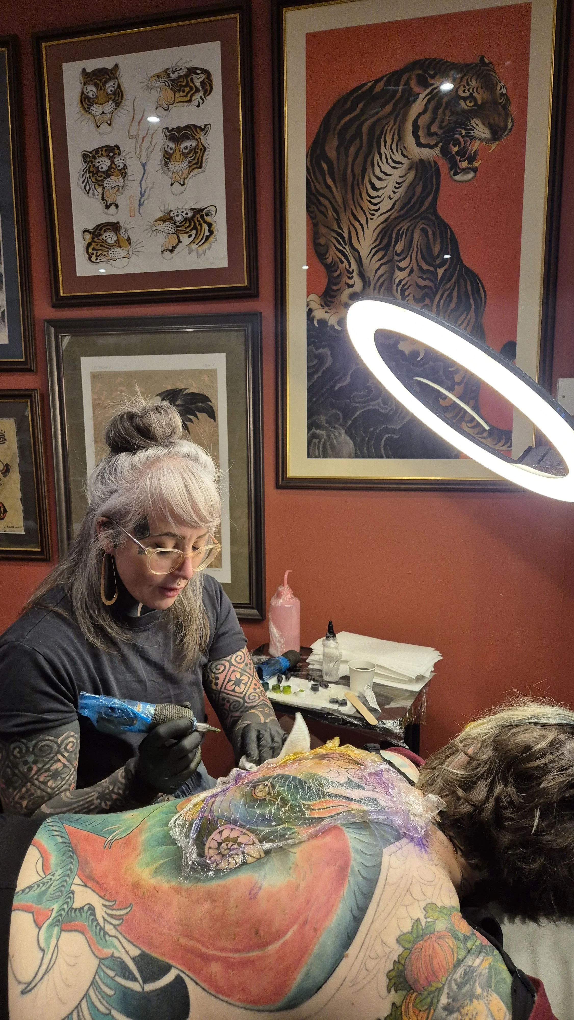 Tattoo artist working on a colorful back tattoo of a dragon, with framed tiger art on the wall behind her and a tattooation ring light illuminating her work.