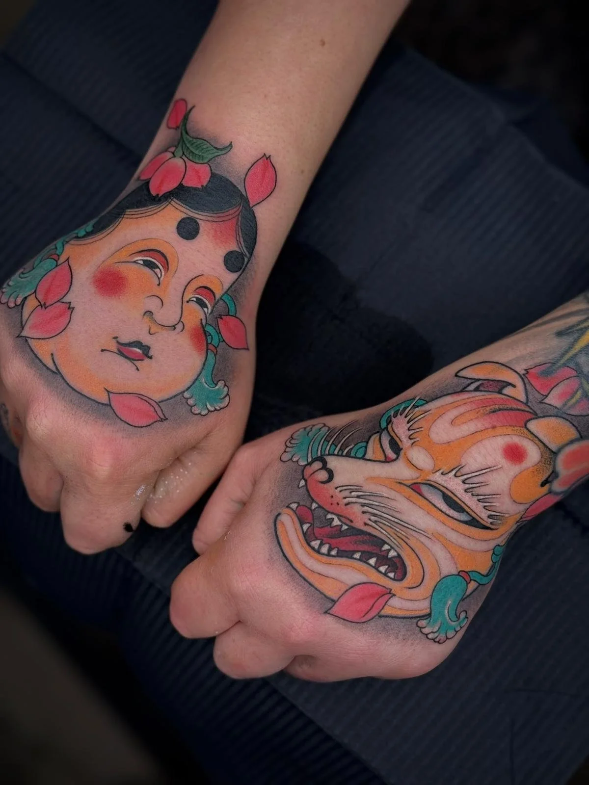 Colorful traditional Japanese style tattoos on hands, featuring a face with a smiling expression and a fierce animal face, both adorned with pink flower petals and teal accents.