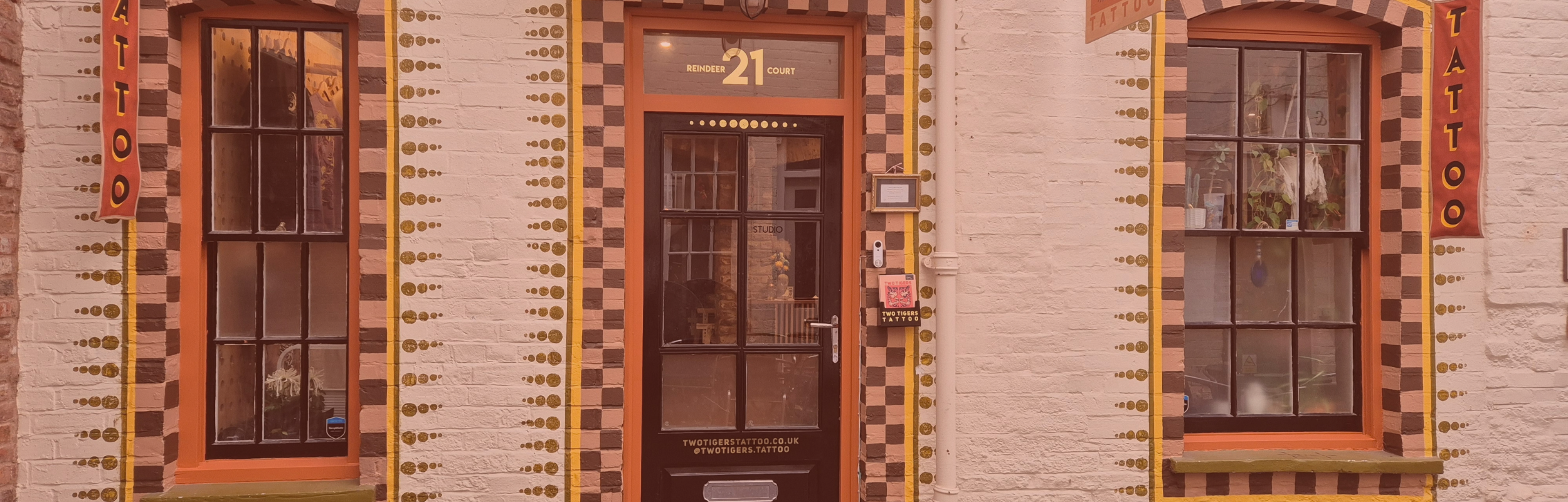 The facade of a tattoo studio with a brick exterior, two large windows, and a glass door painted in orange and decorated with yellow and brown patterns. There are signs indicating it is a tattoo studio, including the website and social media information on the door.
