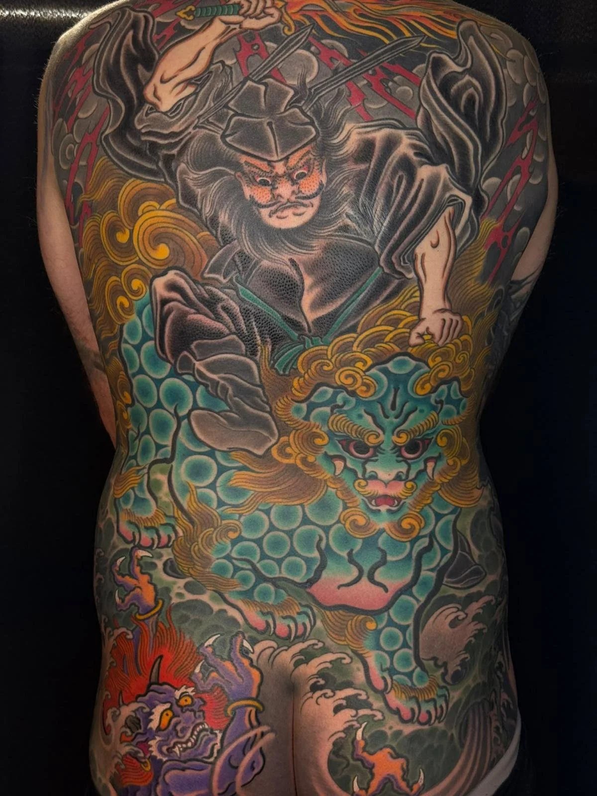 Colorful Japanese-style tattoo covering the back, depicting a fierce lion duel with a tiger, with swirling clouds, flames, and traditional motifs.