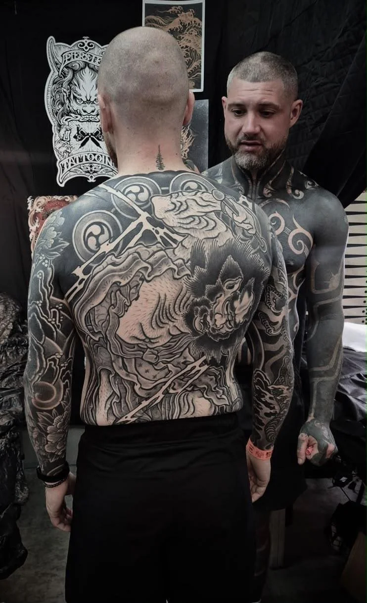 Two men with extensive black and gray tattoos. The man in the foreground has a large dragon tattoo covering his back and arms. The man in the background has tattoos on his neck and chest.