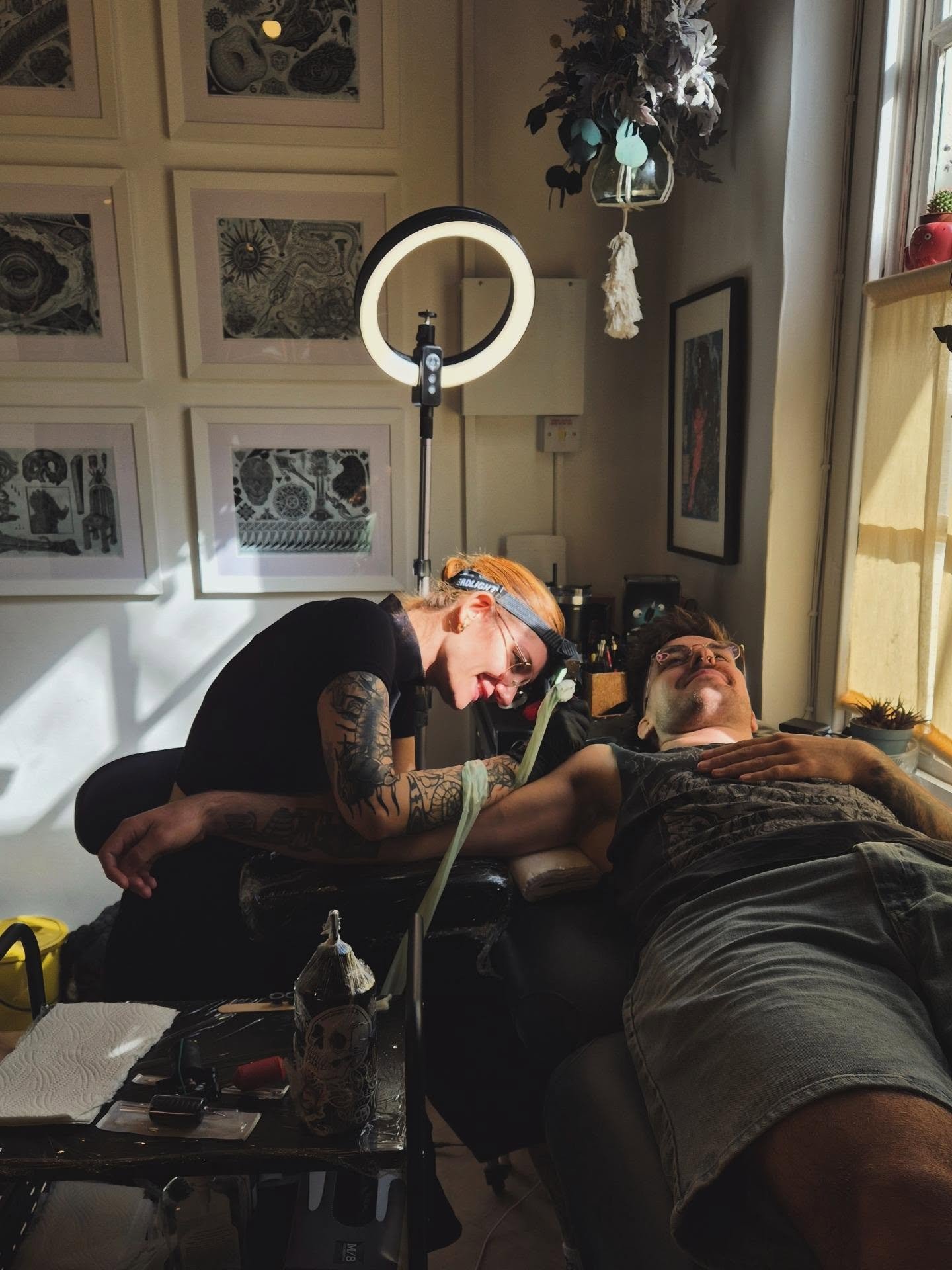 A tattoo artist with tattoos on her arms is giving a tattoo to a man lying on a reclining chair, smiling and relaxed. The artist has orange hair and is wearing glasses. A ring light is positioned above them. The room has framed artwork on the wall, a window with yellow curtains, and various tattoo supplies on the table.