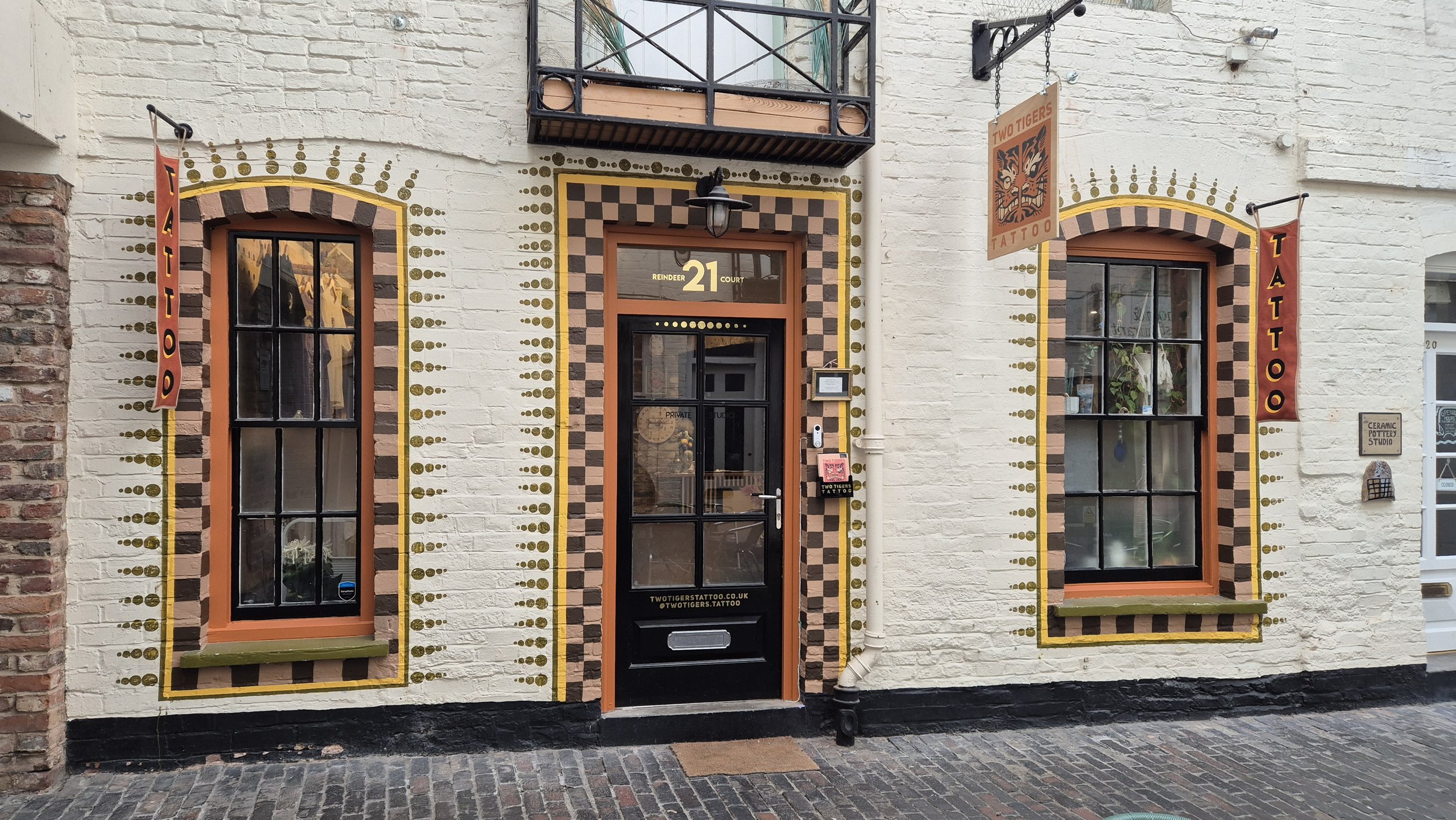 Exterior of a tattoo studio with brick and painted walls, featuring two large arched windows, a door in the center, and decorative paint patterns. Signage includes 'TWO TIGERS TATTOO' banners and a sign reading 'TATTOO' and 'CERAMIC POTTERY STUDIO' n