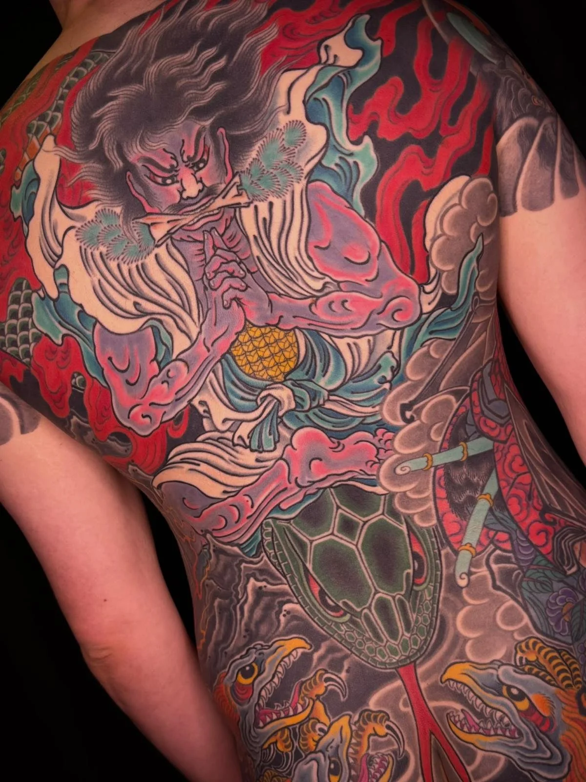 Colorful traditional Japanese style tattoo on a person's chest and arm featuring a fierce demon or ogre holding a smoking pipe, with a green turtle, clouds, and various decorative elements.
