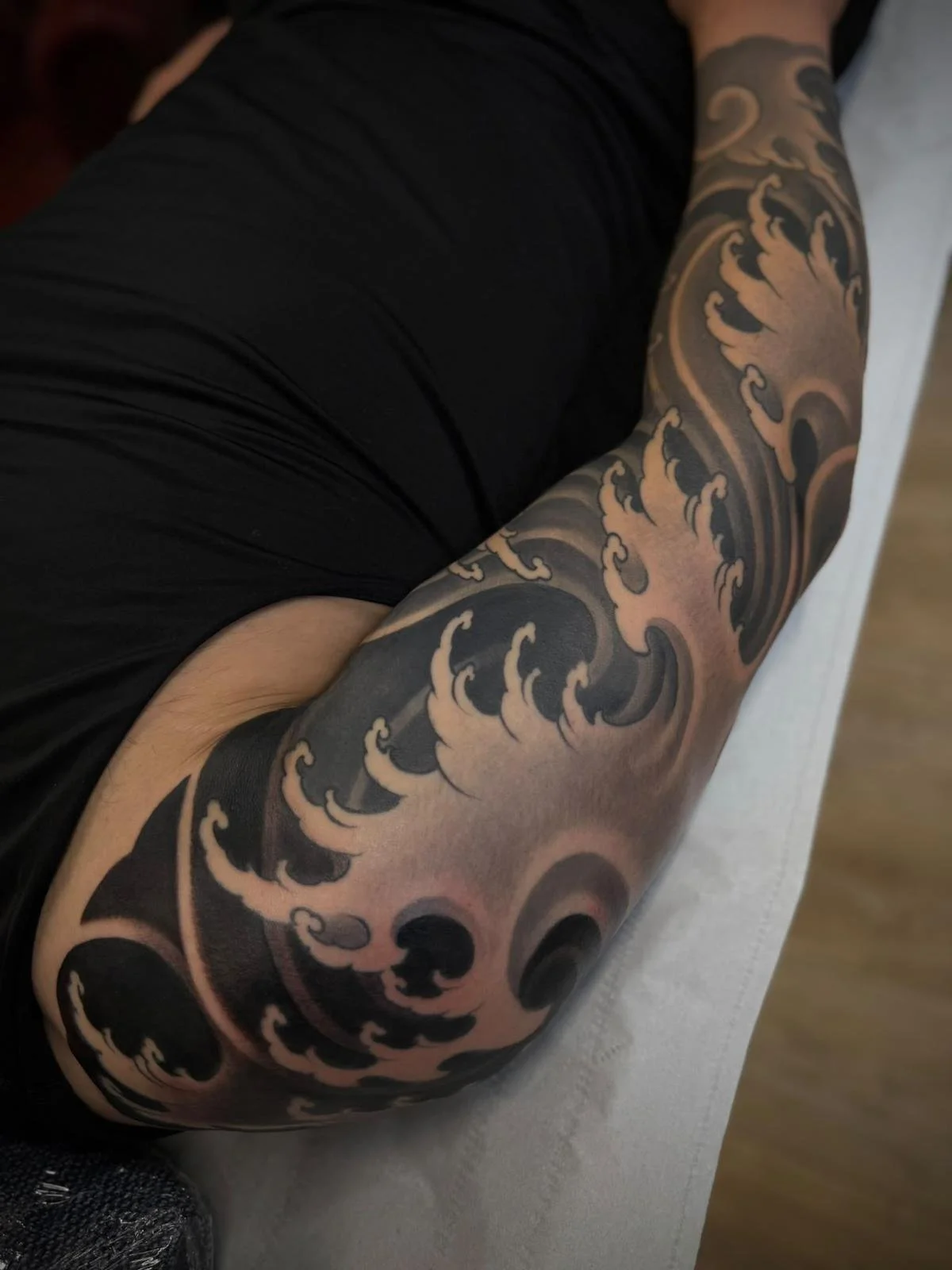 Close-up of a person's arm with a detailed black and gray Japanese-style wave tattoo, wearing a black shirt.