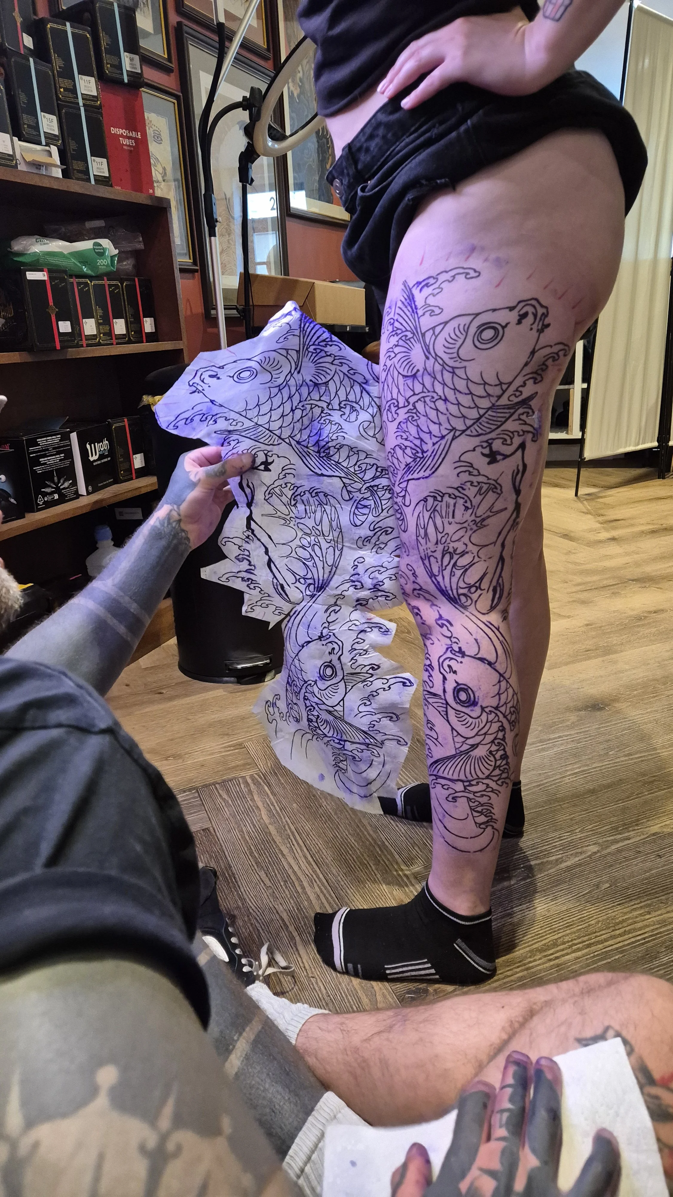 Person with tattooed legs and black sneakers standing as someone prepares to tattoo a koi fish design on their thigh using stencil paper. The tattoo artist's hand is visible holding the stencil paper. The background shows shelves with tattoo supplies
