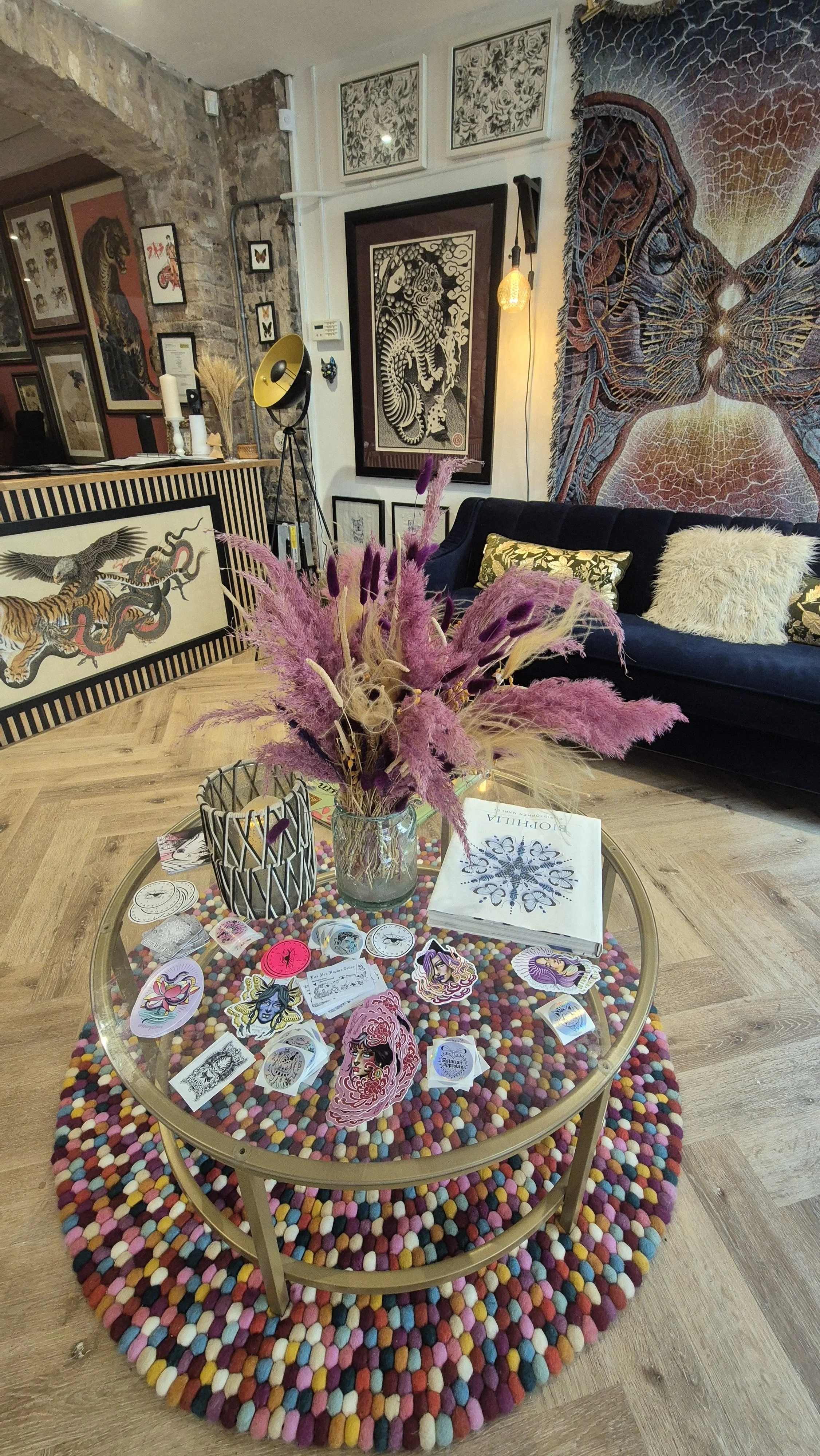 Cozy interior with artwork and decor, including a glass coffee table with stickers and illustrations, a colorful circular rug, a black sofa with pillows, a bouquet of pink pampas grass, and various artworks on the walls.