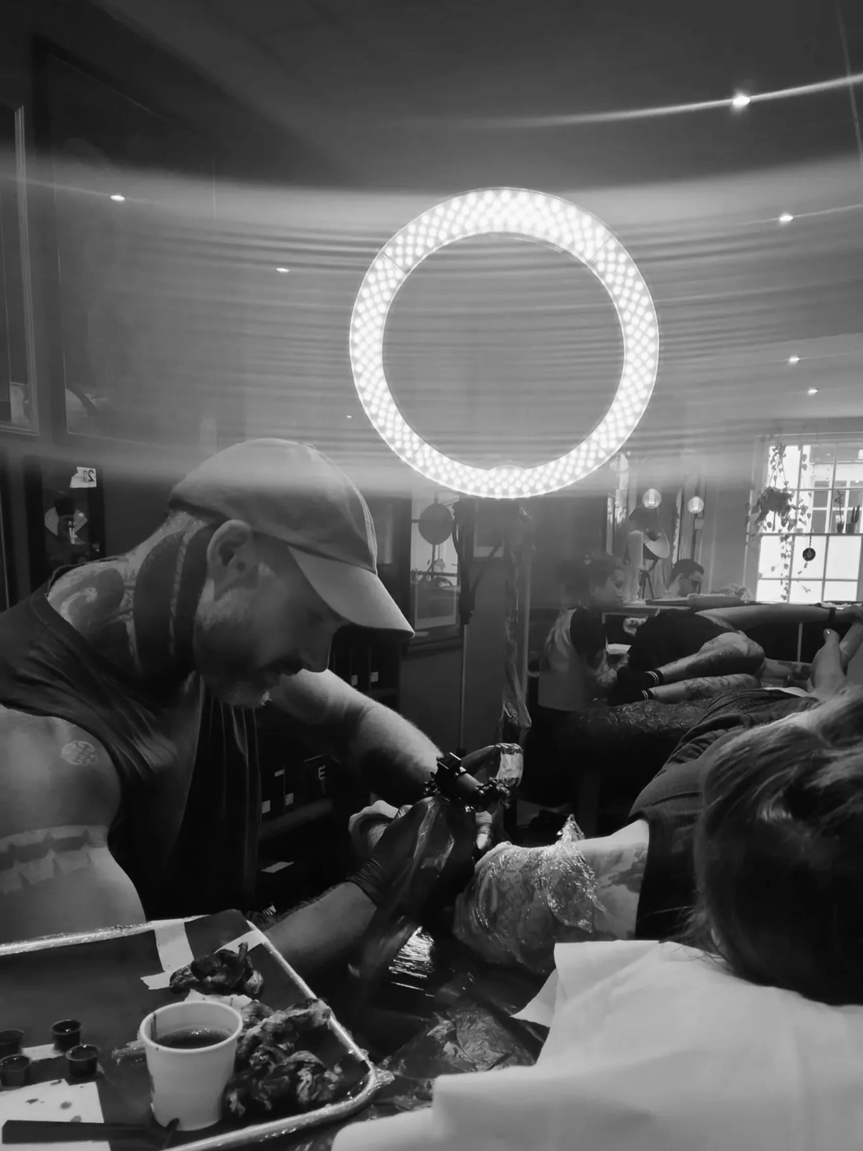 A black and white photo of a person wearing a mask and cap, working with their hands at a table with food and drinks, possibly in a restaurant or cafe, with other people visible in the background.