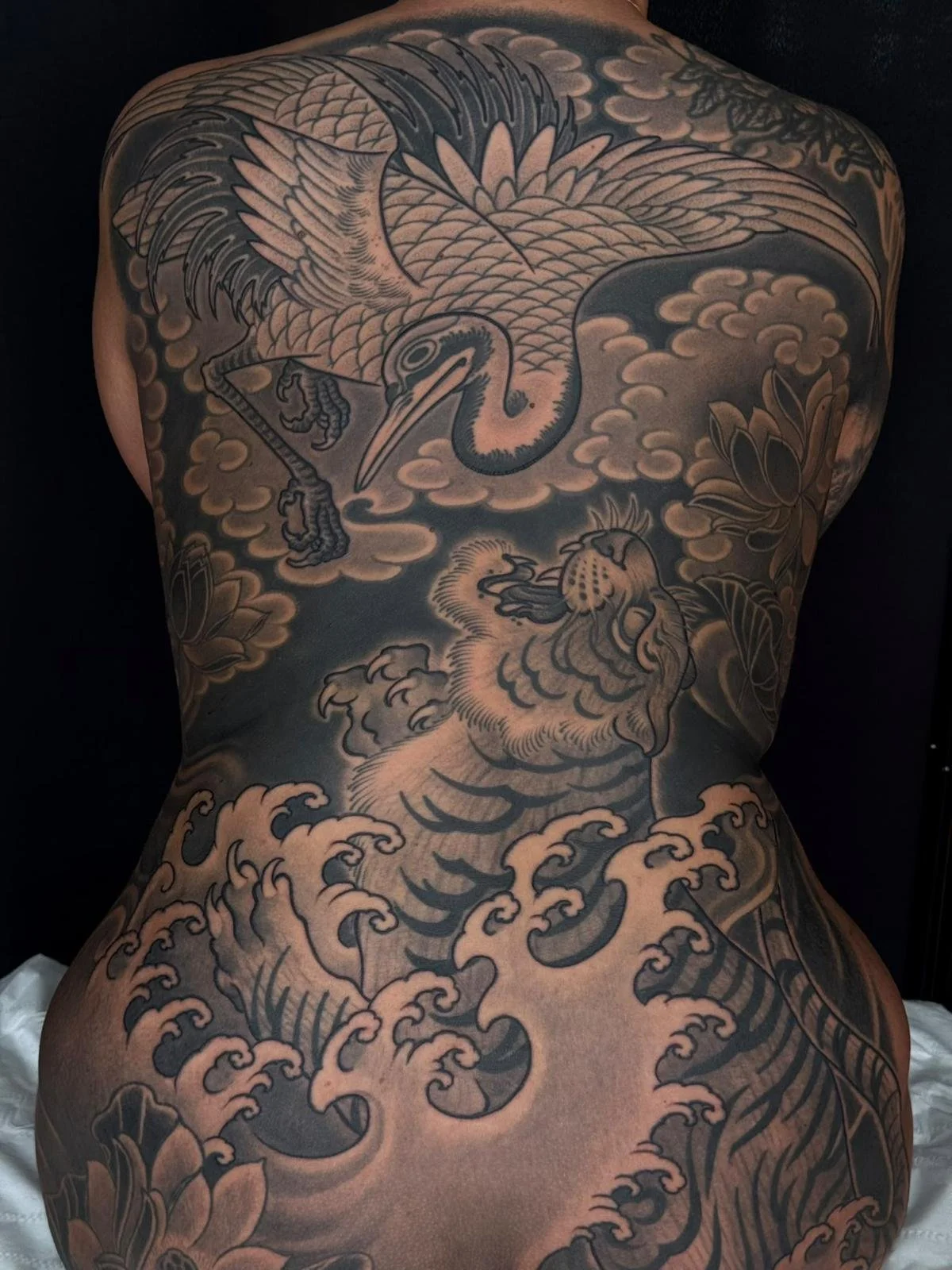 A back tattoo of a large bird, possibly a crane or heron, flying over stylized waves and clouds, with dragons or other mythical creatures around it.