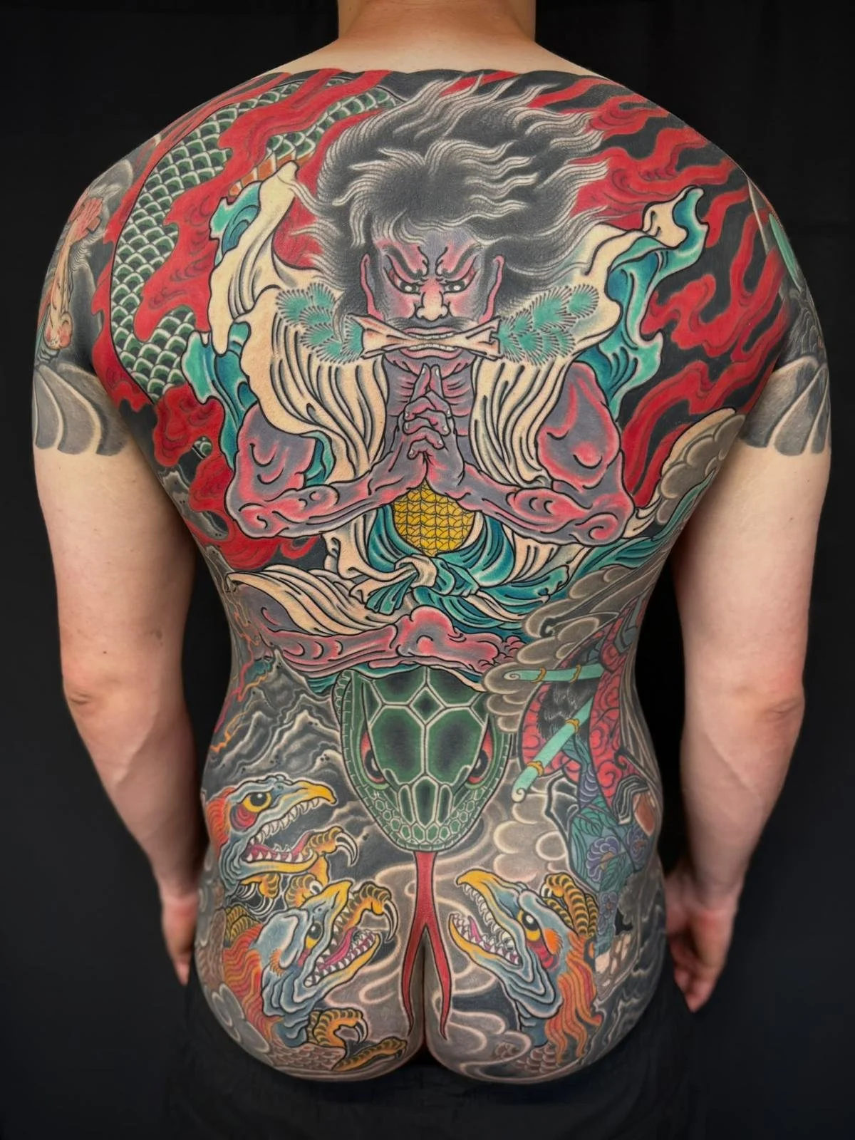 Colorful full-back Japanese style tattoo featuring a fierce figure with wild hair and a mustache, holding a leafy branch with a sacred halo, surrounded by swirling clouds, dragons, turtles, and traditional Japanese motifs.