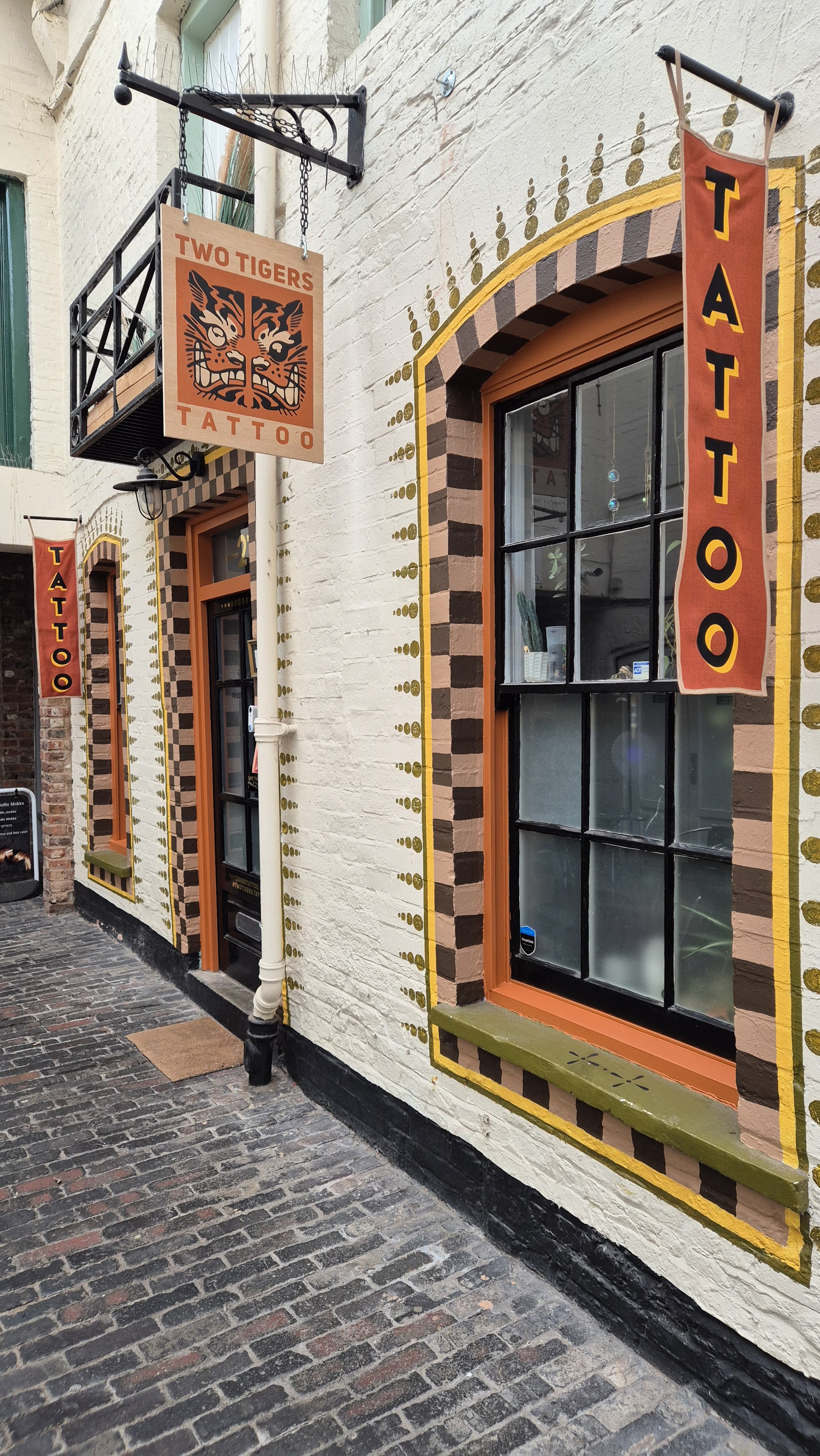 Exterior of a tattoo shop with colorful painted window and door frames, a vertical banner reading 'TATTOO', and a hanging sign with a tiger face and the words 'TWO TIGERS TATTOO'