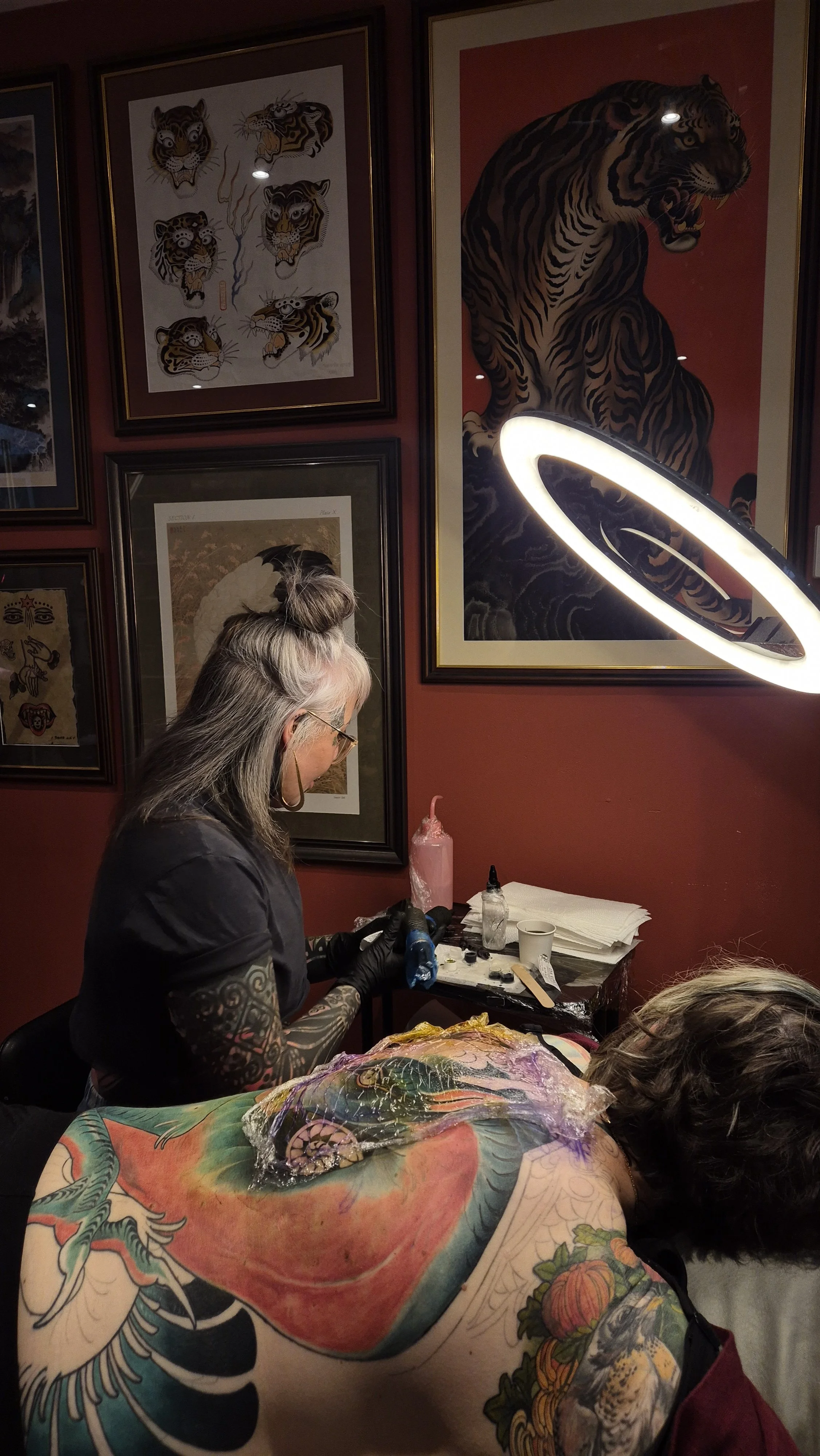 Tattoo artist working on a person's tattoo in a room decorated with tiger artwork and framed art on the wall.