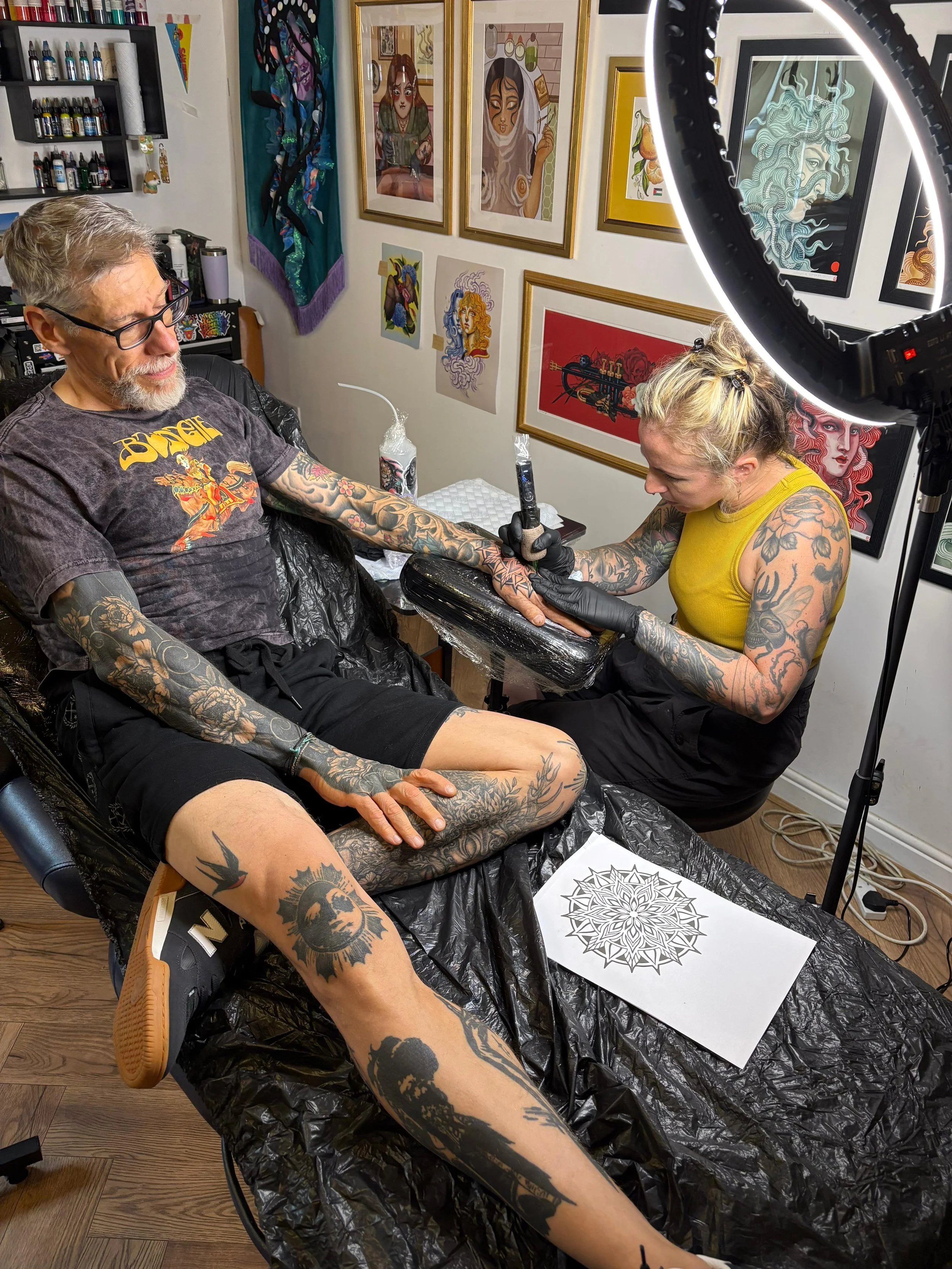 A tattoo artist is tattooing a man's arm in a tattoo studio. The man has tattoos on his arms and legs and is seated on a chair with a black plastic covering. The tattoo artist, with tattoos on her arms, is wearing a yellow tank top and black gloves, 