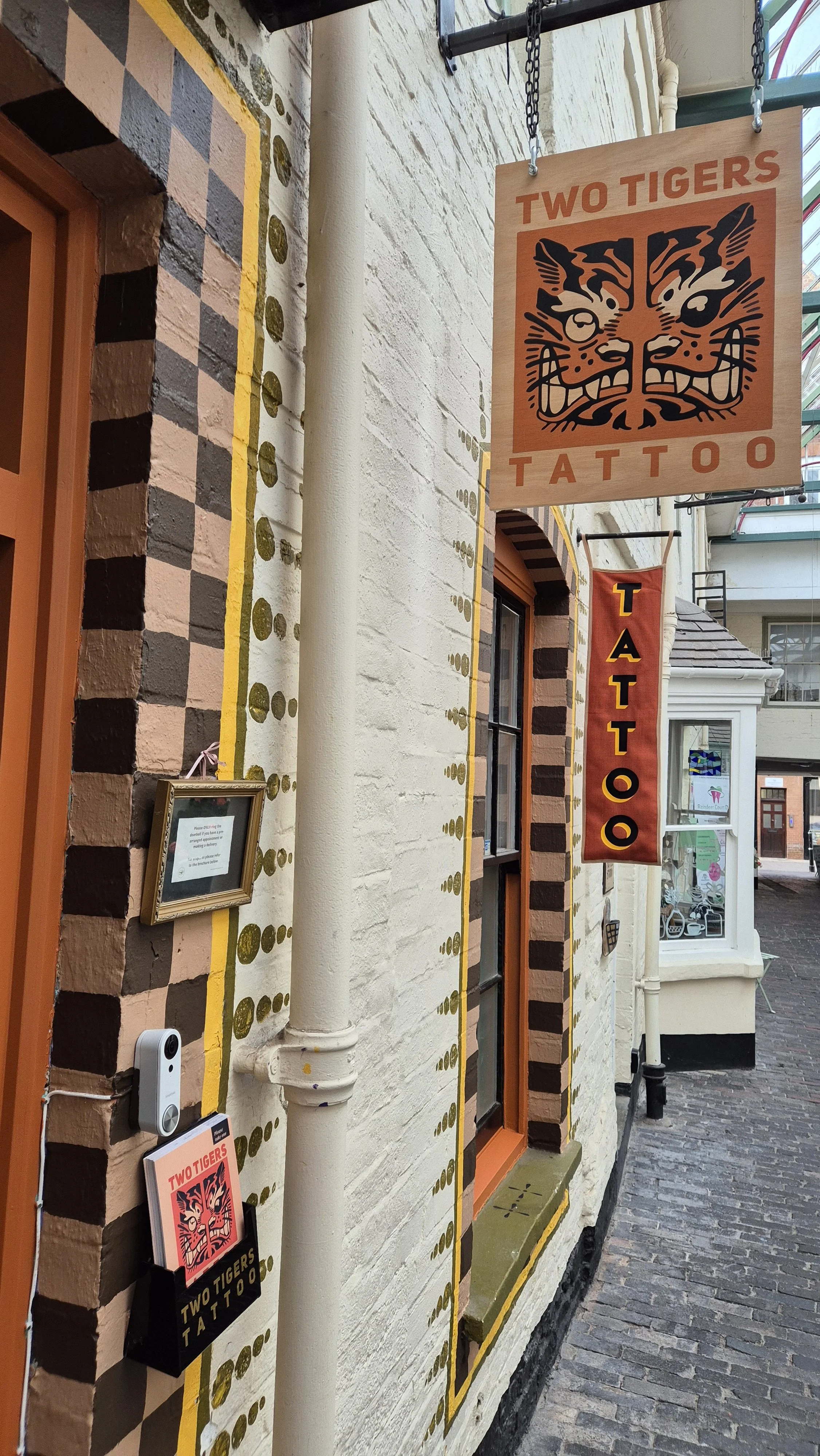 Exterior of a tattoo studio with decorative wall details, a sign with a tiger face logo reading "Two Tigers Tattoo," and a vertical banner with the same logo and text.