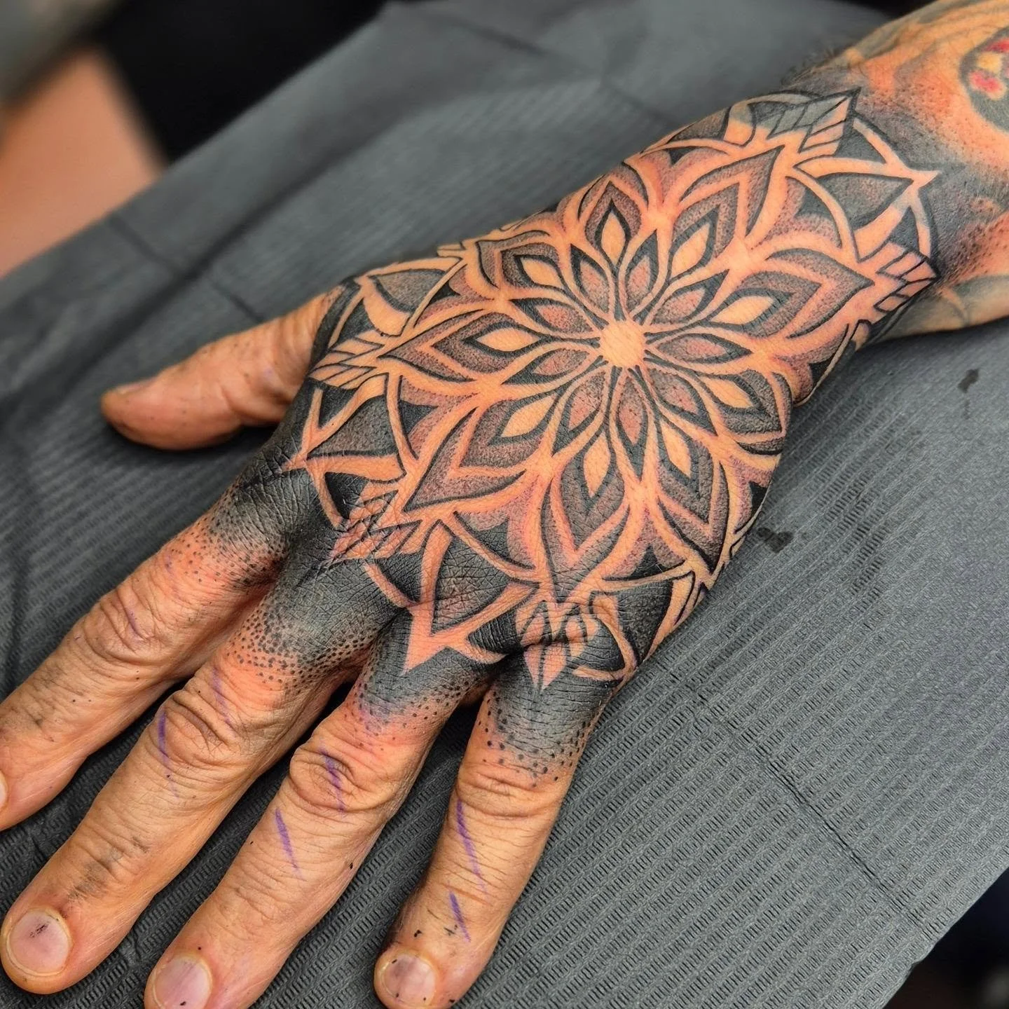 A person's hand with tattooed fingers and a large geometric tattoo on the back of the hand featuring a complex pattern of black outlines and orange shading.