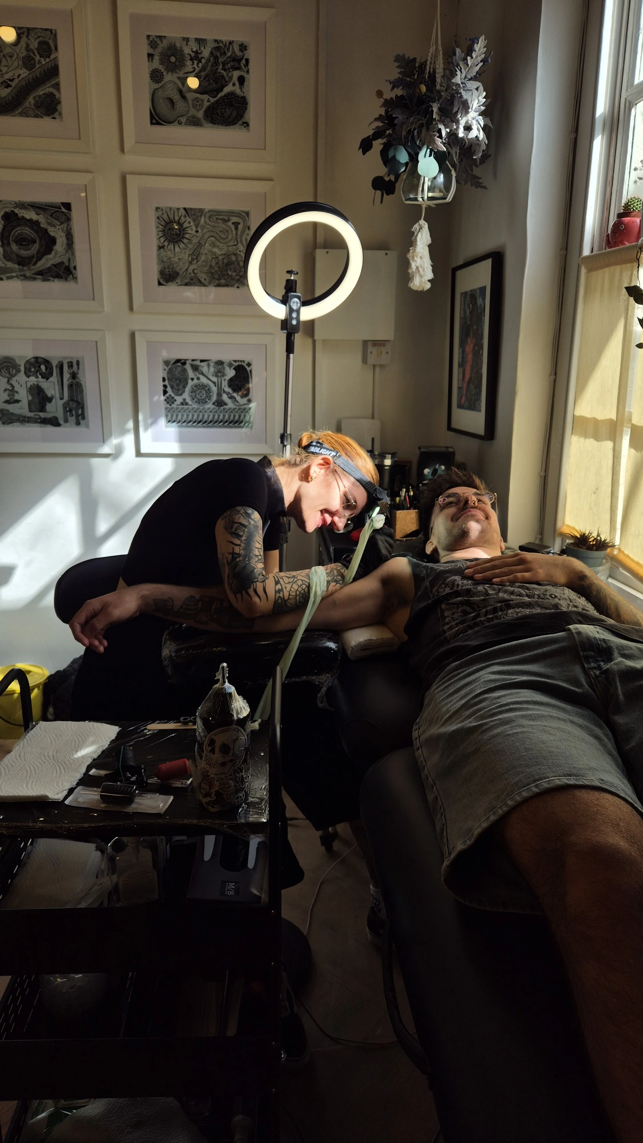 Tattoo artist with orange hair and glasses getting a tattoo on a man's arm in a well-lit studio. The man is lying on a chair, looking relaxed. Artwork hangs on the walls, and tattoo equipment is on a nearby table.