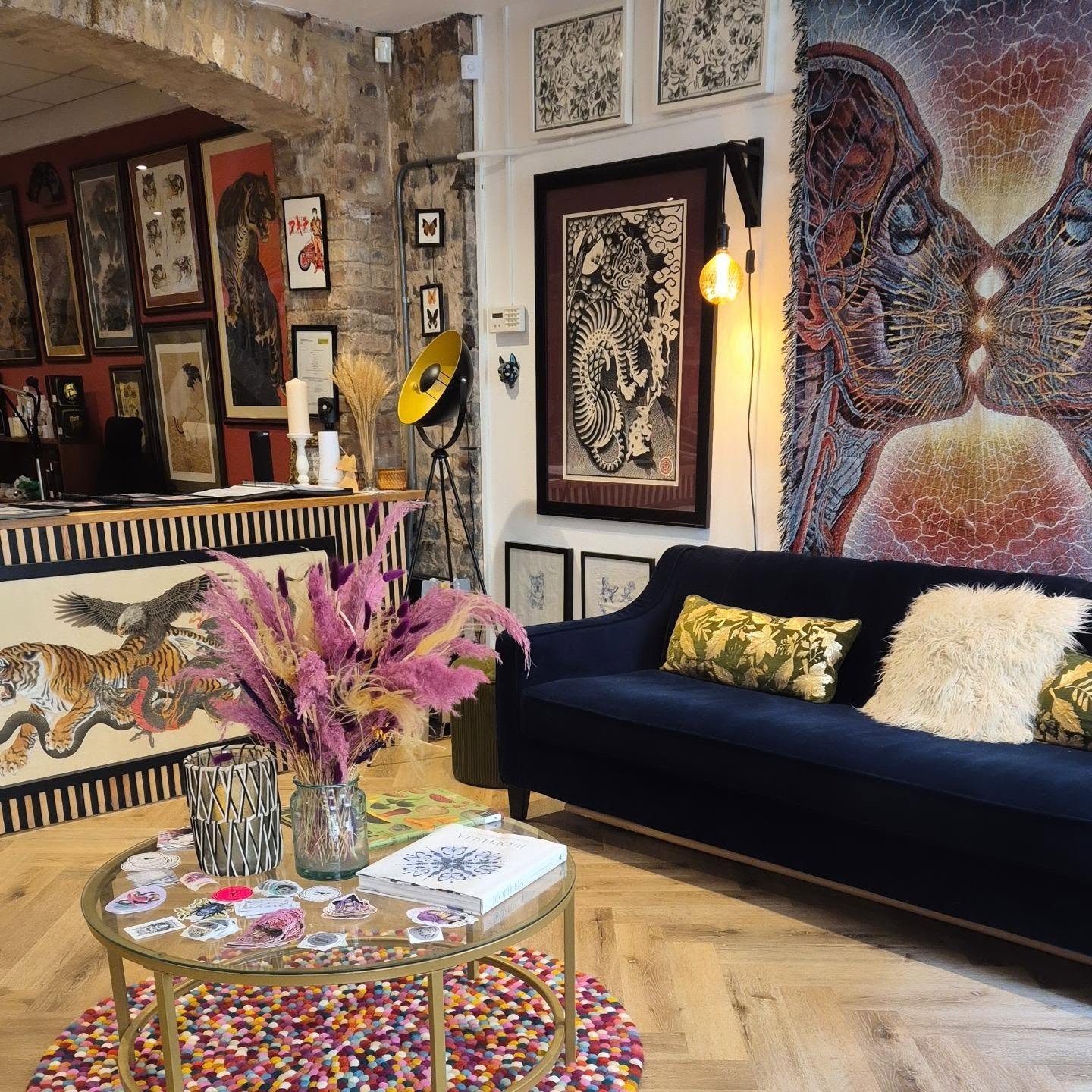 A living room with a black velvet sofa, a colorful circular rug, and a glass-topped coffee table with magazines, stickers, and a vase of pink and yellow dried flowers. The walls are decorated with various framed artworks, including a large tapestry o