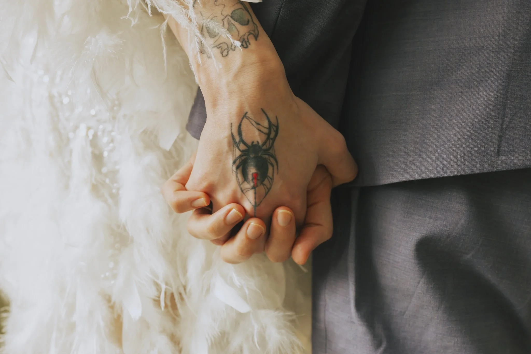 Two hands intertwined, with visible tattoos on one wrist and hand, resting on a gray fabric surface. The background features soft white feathers and textured white fabric.