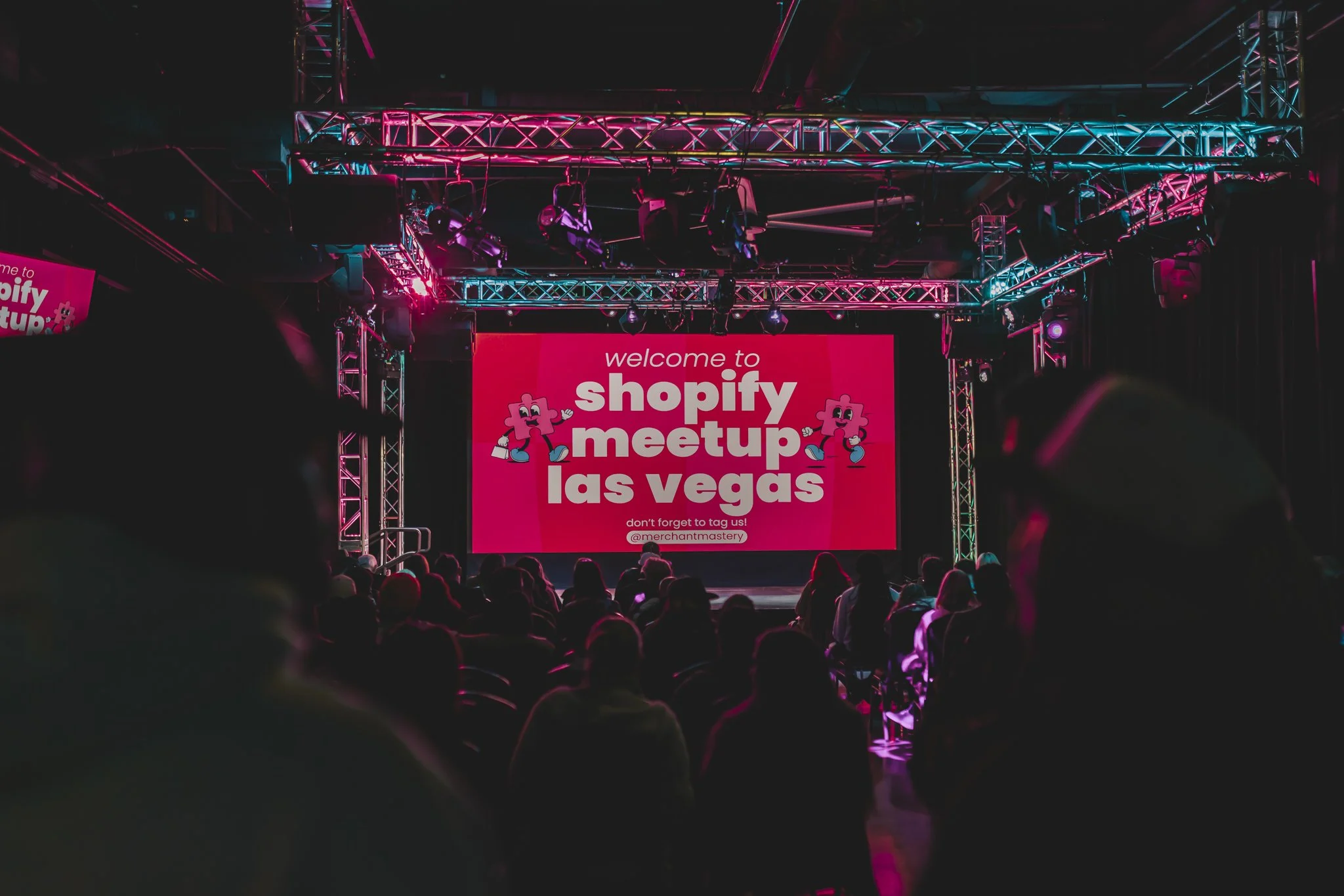 A large digital screen with the text 'welcome to shopify meetup las vegas' and two cartoon graphics of a pink puzzle piece character. The stage is surrounded by metal trusses with colorful lighting, and a crowd of people is visible watching the event.