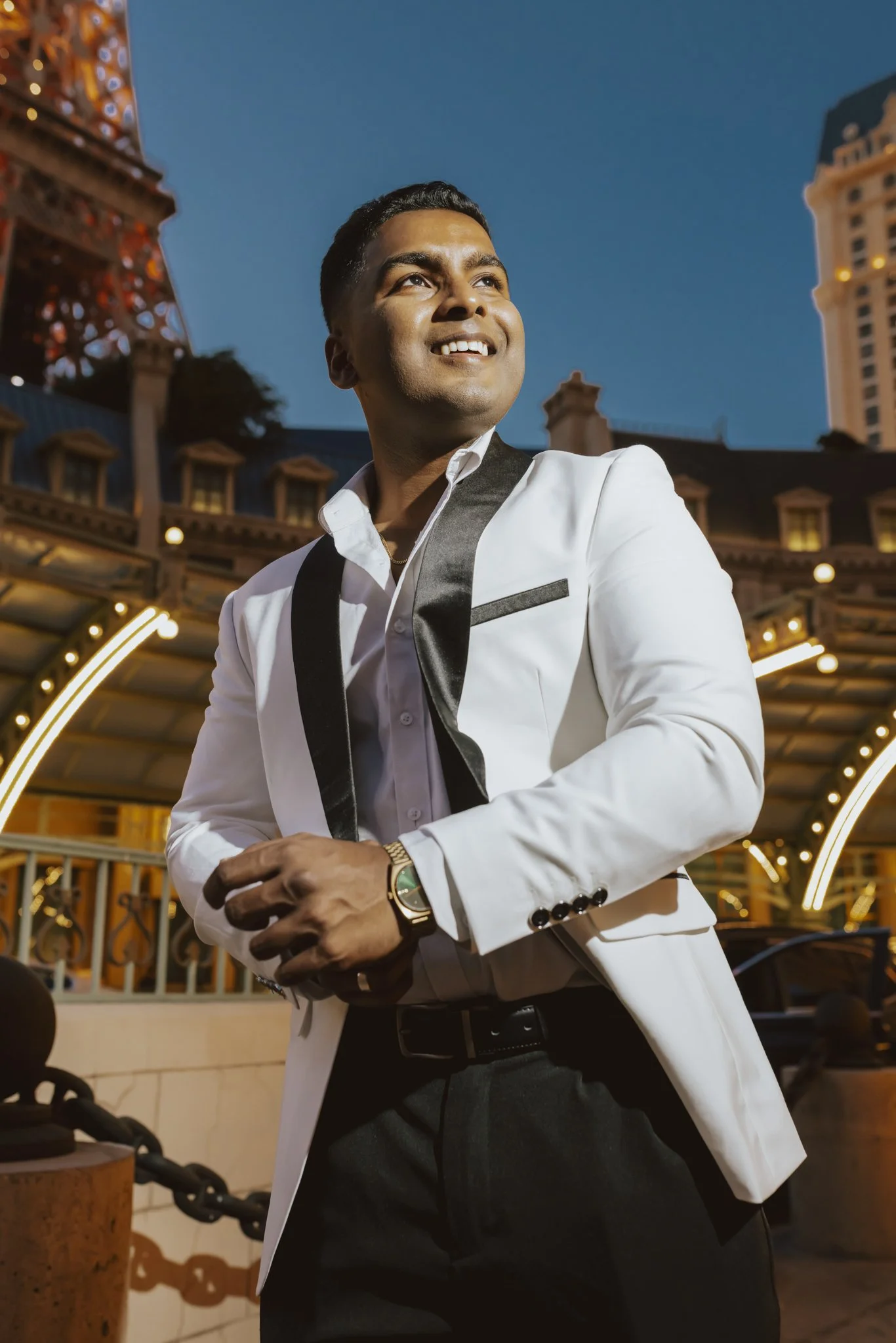 A man dressed in a white tuxedo jacket with black lapels and black pants, standing outdoors at night, with a clear sky and a decorative building illuminated with lights in the background.