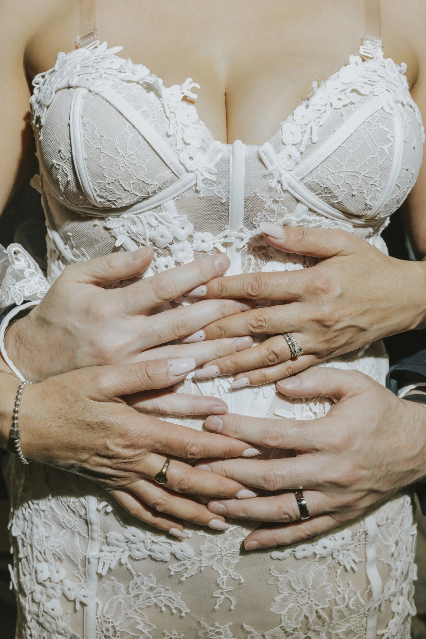 Close-up of a woman wearing an ivory, lace, and embroidered bustier, with hands gently placed on her stomach, including rings and nail polish.