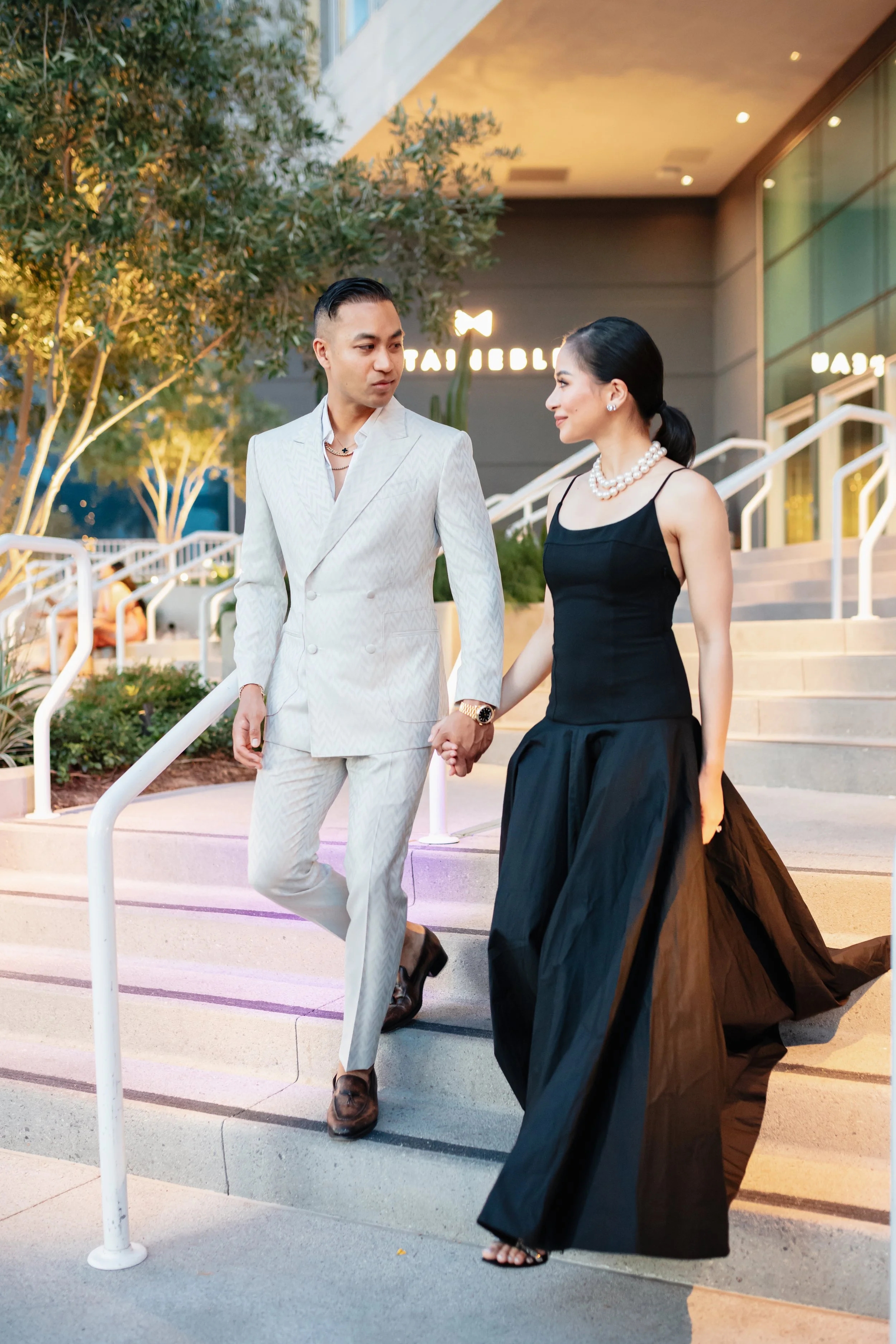 A man and woman dressed in formal attire holding hands and walking down stairs outside a modern building during evening. The man is wearing a white suit and brown shoes, and the woman is wearing a black gown with pearls and black sandals. They are looking at each other with a cityscape background featuring illuminated signs and greenery.