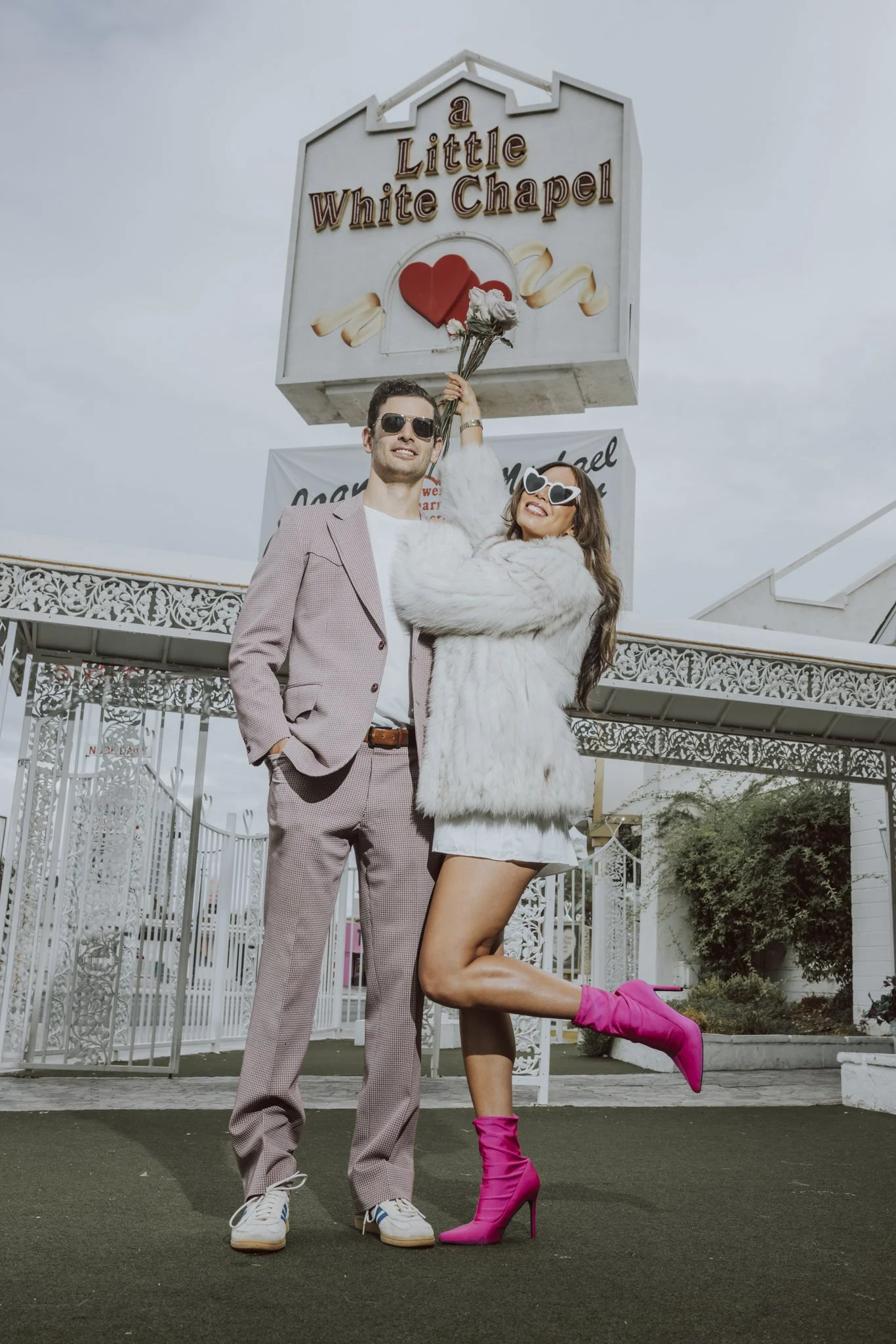 Two people posing in front of the sign for 'A Little White Chapel.' The person on the left is a man dressed in a light pink checked suit, white shirt, white sneakers, and sunglasses, standing with one hand in his pocket. The person on the right is a woman in a white fur coat, black shorts, bright pink high-heeled boots, and white sunglasses with heart-shaped lenses. She is holding flowers above her head and lifting one leg, showing a playful pose.