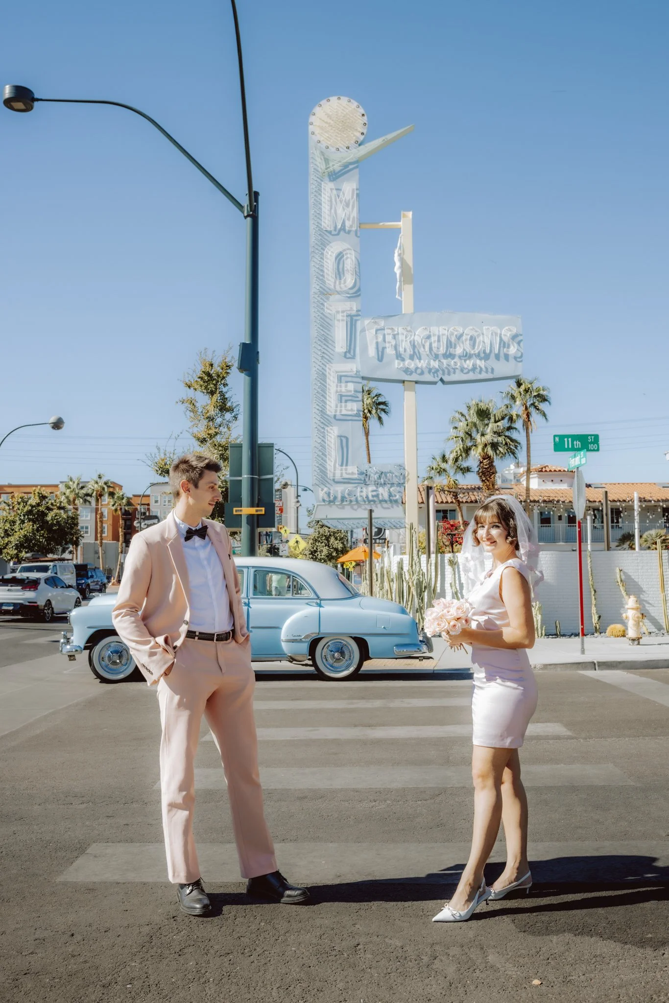 A bride in a white dress holding a bouquet of pink roses stands in a crosswalk while a groom in a light pink suit with a black bow tie stands nearby on a sunny day. There is a classic car and palm trees in the background, with a big vertical sign that reads 'MOTEL' and other buildings and street signs.