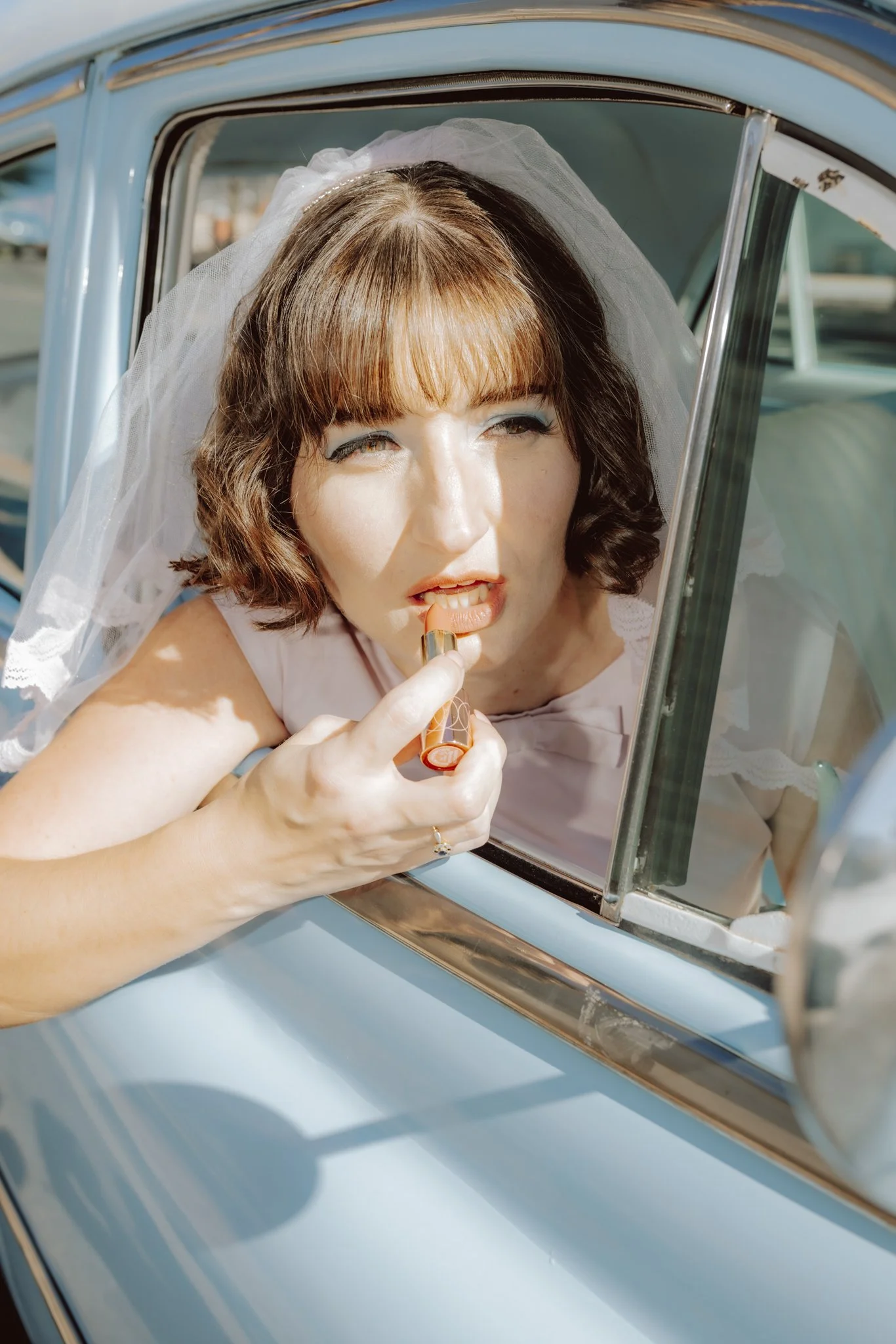 A woman with short brown hair and bangs, wearing makeup and a white outfit, is looking out of a classic blue car's window while applying lipstick.