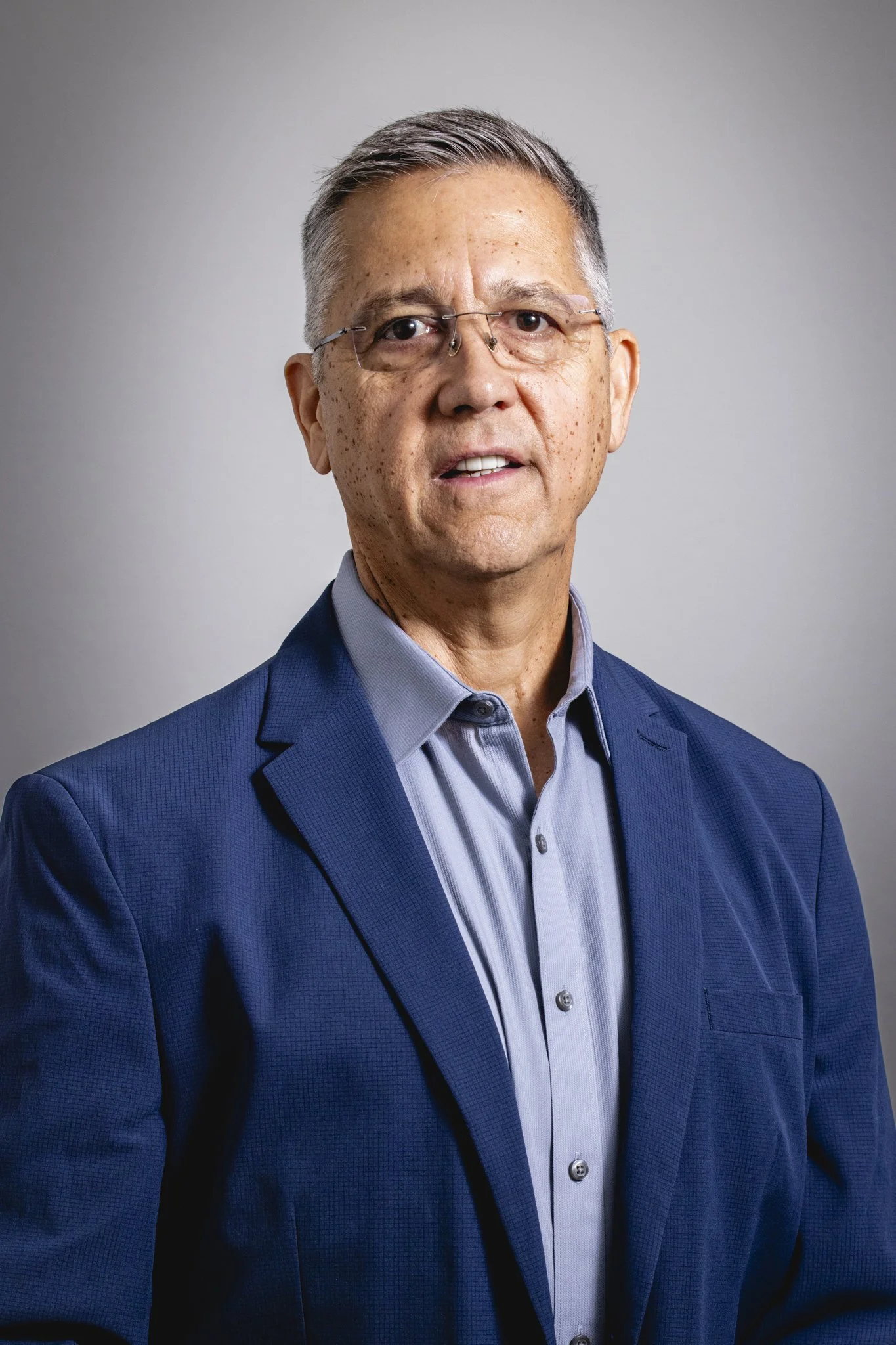 A middle-aged man with gray hair and glasses wearing a blue suit jacket and a light blue dress shirt, standing against a plain gray background.