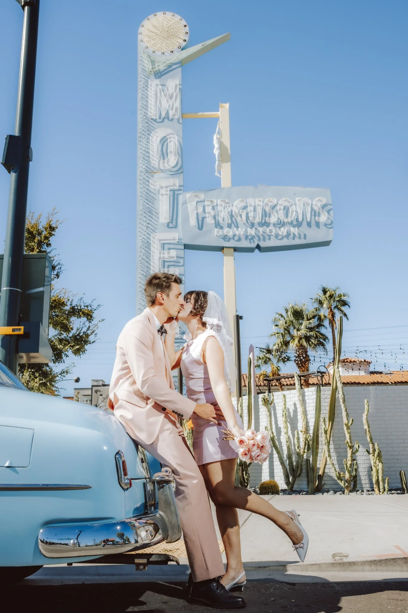 A couple dressed in wedding attire sharing a kiss near a vintage light blue car with a bouquet of pink roses, standing in front of a sign that reads 'Fergusons Downtown' against a clear blue sky.