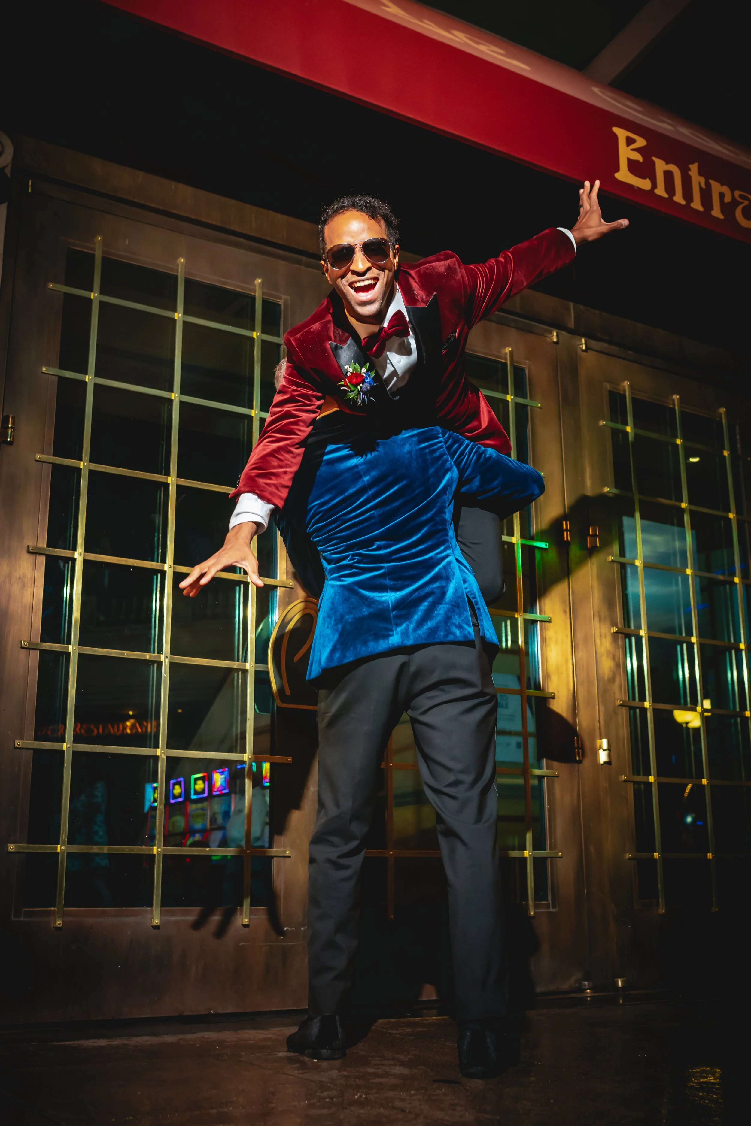 A man wearing sunglasses, a red velvet tuxedo jacket, a bow tie, and gray pants is dancing in front of a glass door with gold accents at night. He has one arm extended and is smiling broadly.