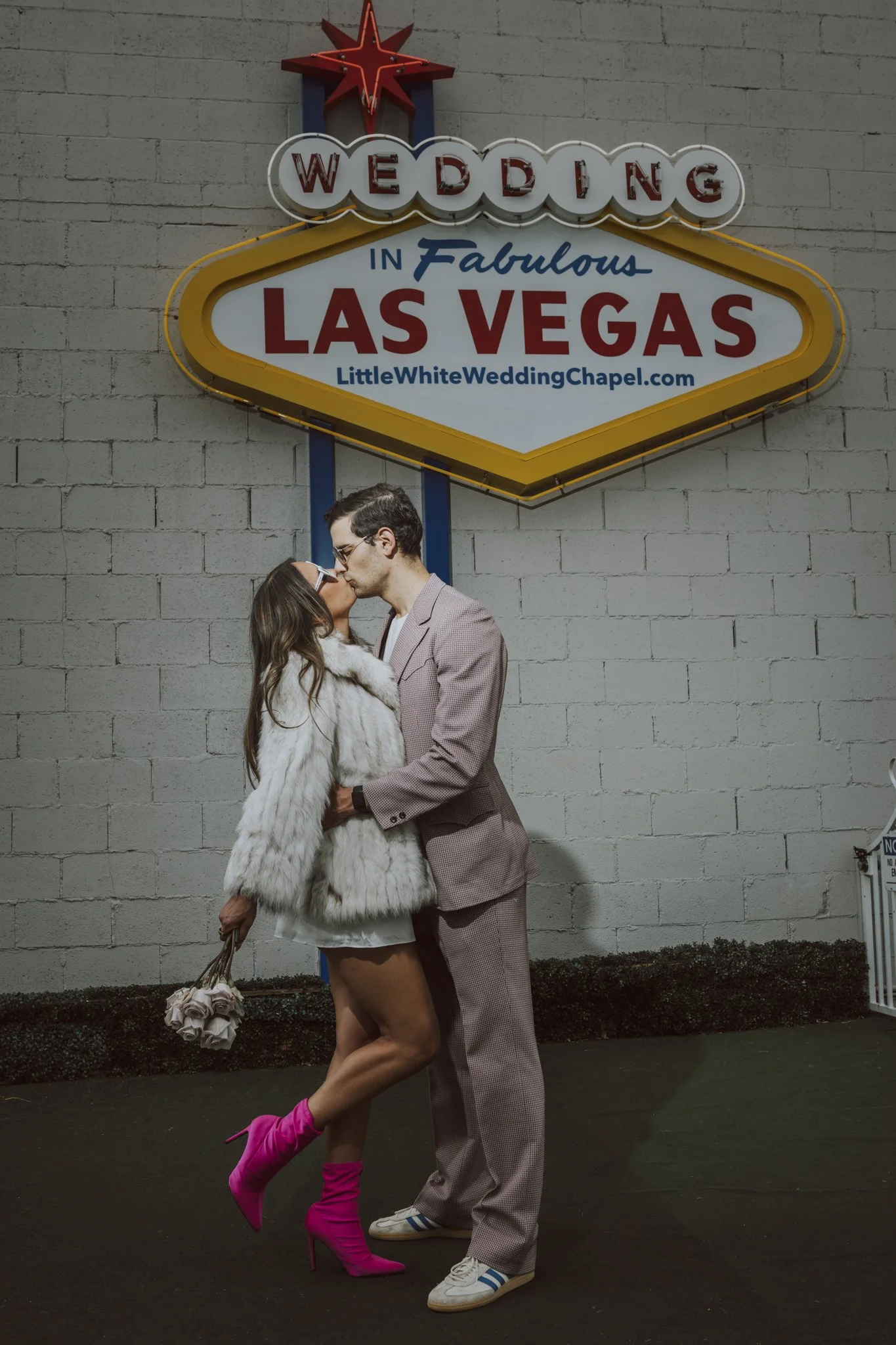 A woman and a man are kissing in front of a sign that reads 'Wedding in Fabulous Las Vegas' on a gray brick wall. The woman is wearing a white fur coat, pink thigh-high boots, and holding a bouquet, while the man is dressed in a light-colored suit and sneakers.
