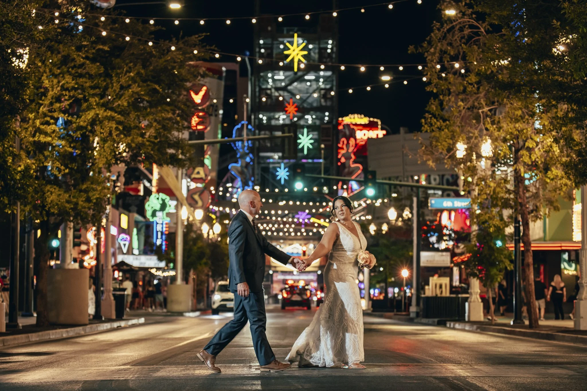 A bride and groom hold hands as they walk across a street at night, illuminated by colorful decorative lights and streetlights, with trees on either side and buildings in the background.