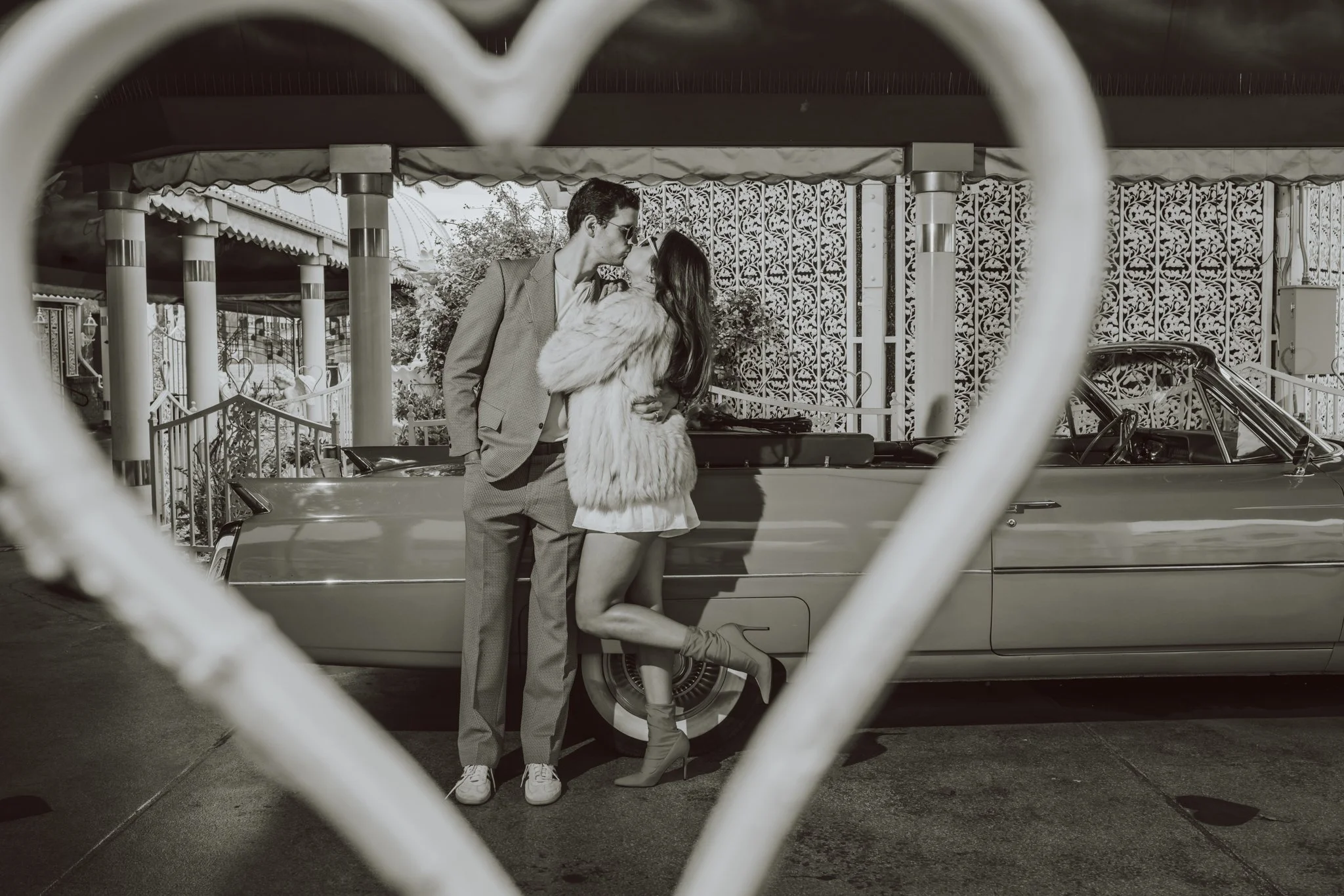 A man and woman sharing a kiss in front of a vintage car, outdoors, behind a decorative metal fence, with the photo framed by a chain-link fence in the foreground.