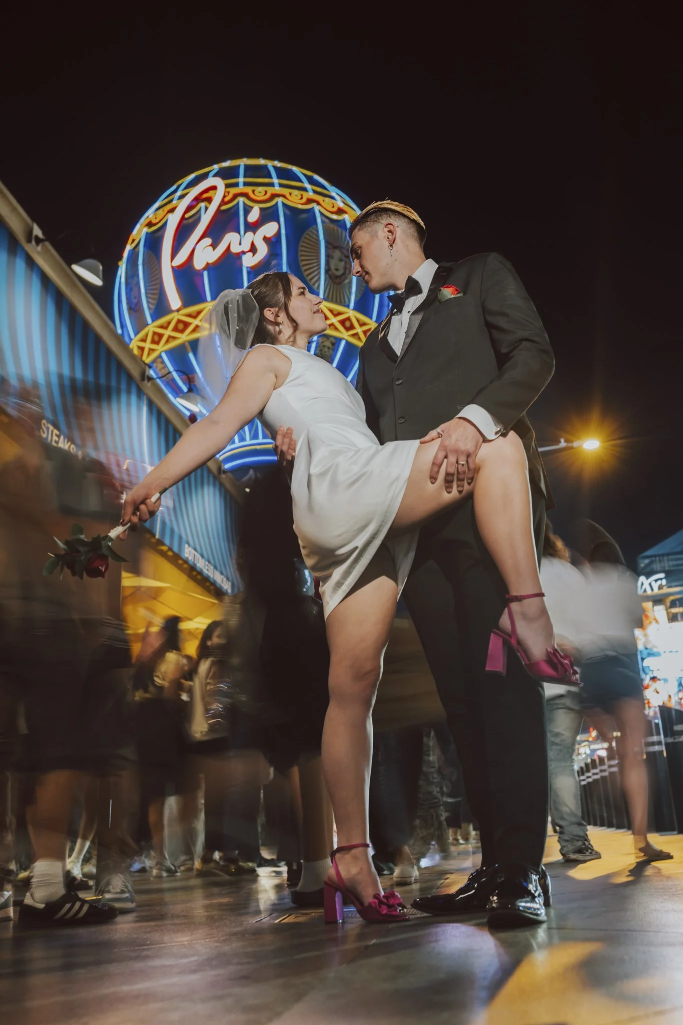 A couple dressed in wedding attire sharing an intimate moment at a fairground with bright neon lights and crowds around.
