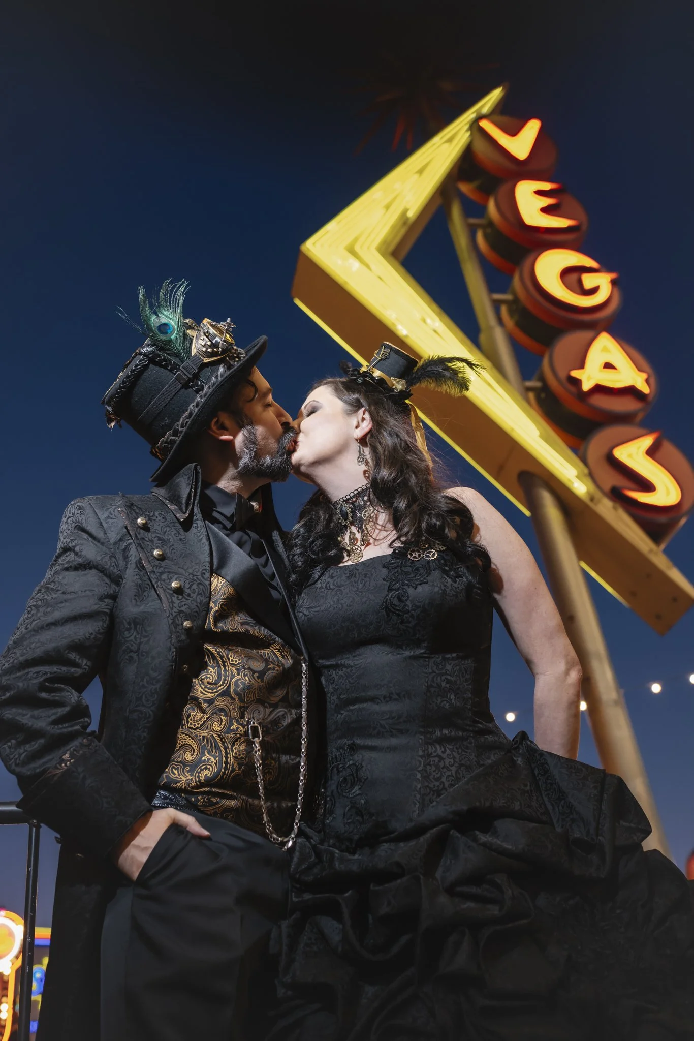 A couple dressed in steampunk-inspired costumes kissing in front of a neon 'Levage' sign at night.