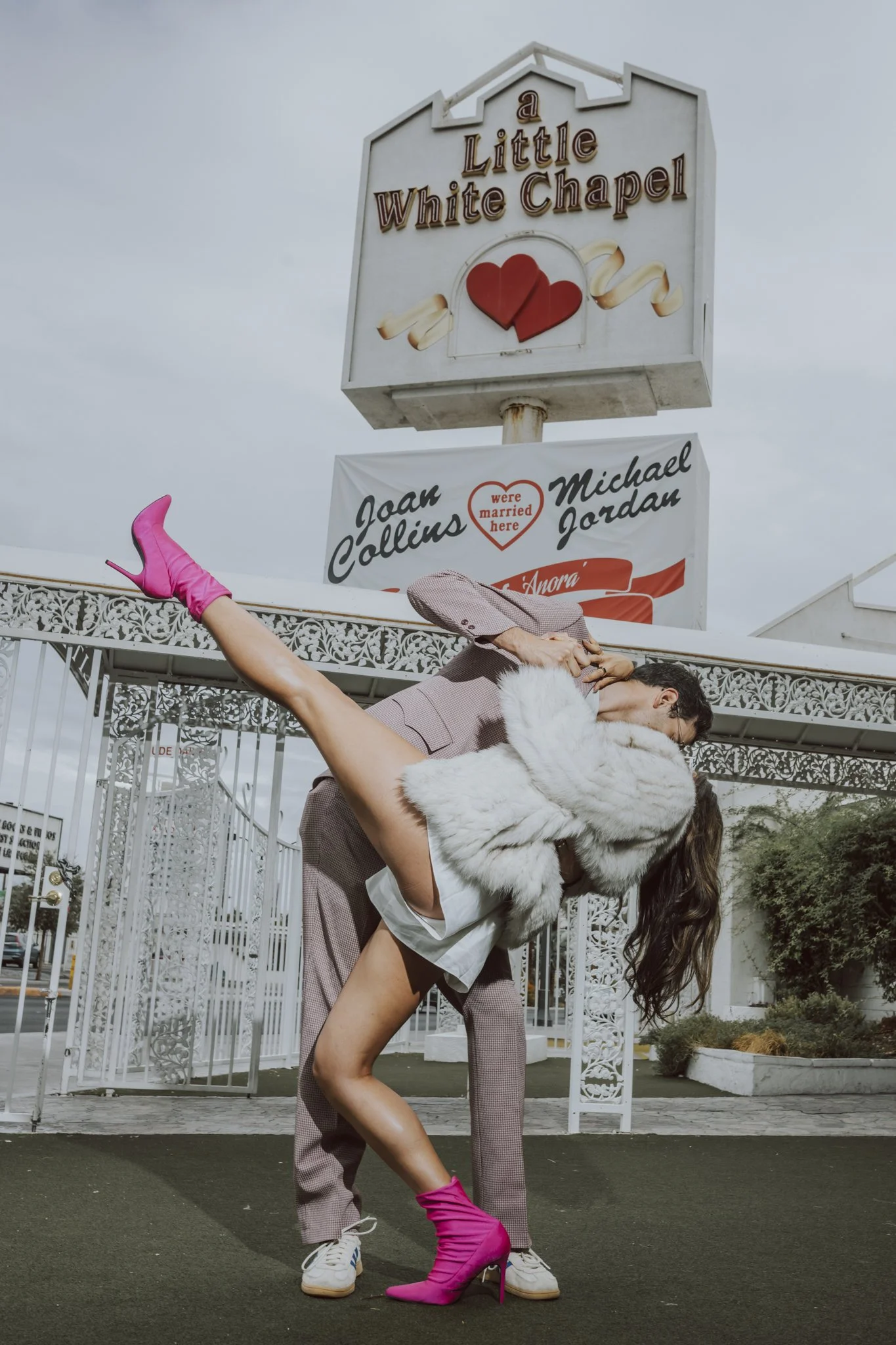 A man in a tan suit with white sneakers is holding a woman with long dark hair, wearing a white fur coat and bright pink high-heeled boots, in front of the sign for a Little White Chapel with red hearts on it.