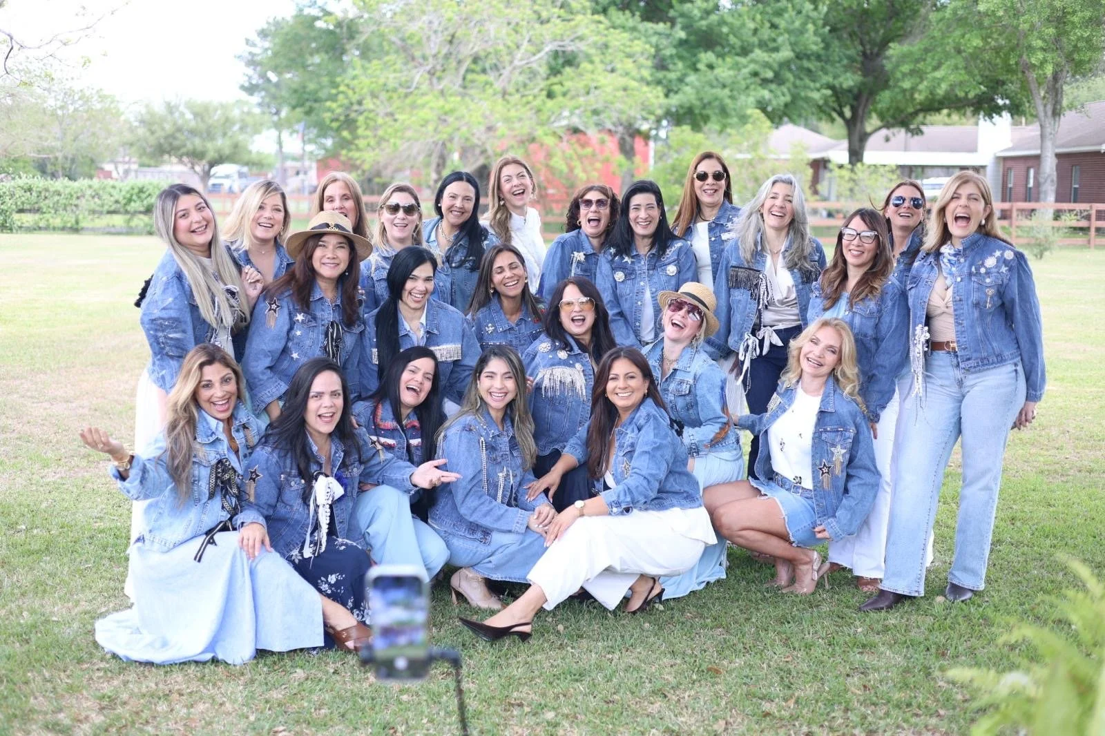 A group of women in denim jackets and pants, smiling and laughing outdoors on a grassy field with trees and houses in the background.
