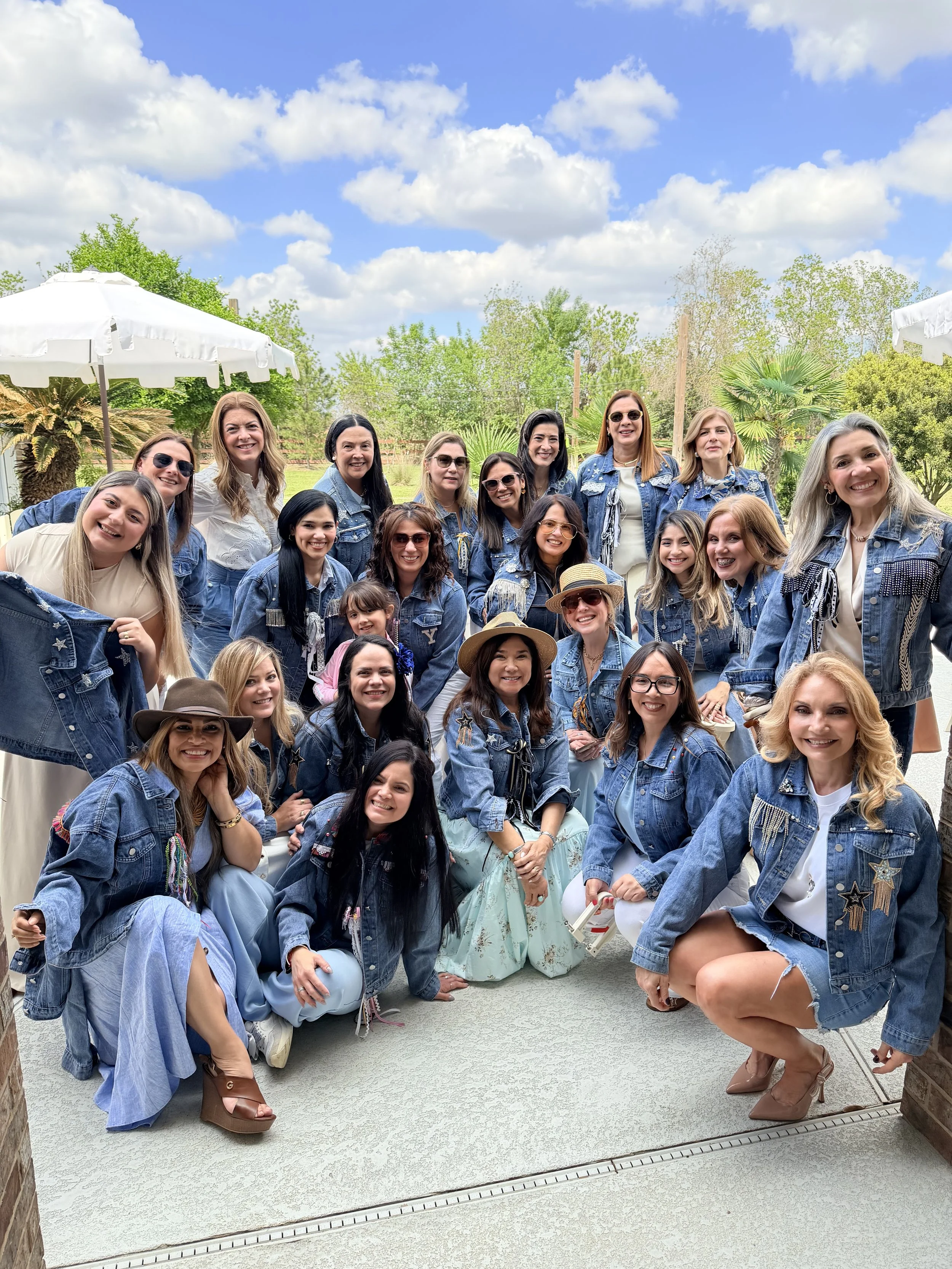 Group of women gathered outdoors, all wearing denim jackets and smiling for a photo under blue skies with clouds, with greenery and trees in the background.