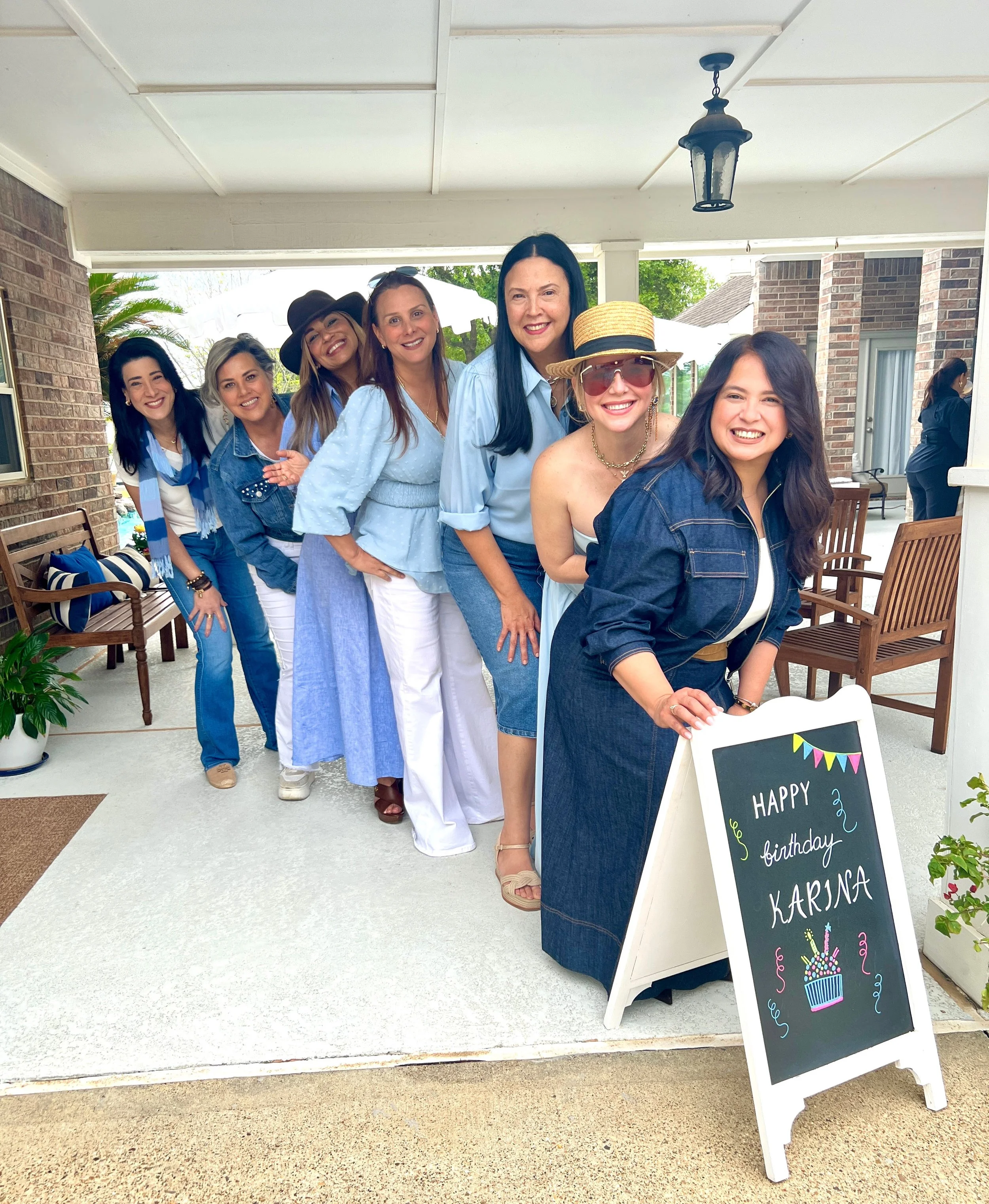 A group of women standing in line on a porch, celebrating Karina's birthday. They are smiling, dressed casually in blue and white, with some wearing hats and sunglasses. A chalkboard sign in front says, 'Happy birthday Karina' with a drawing of a bir