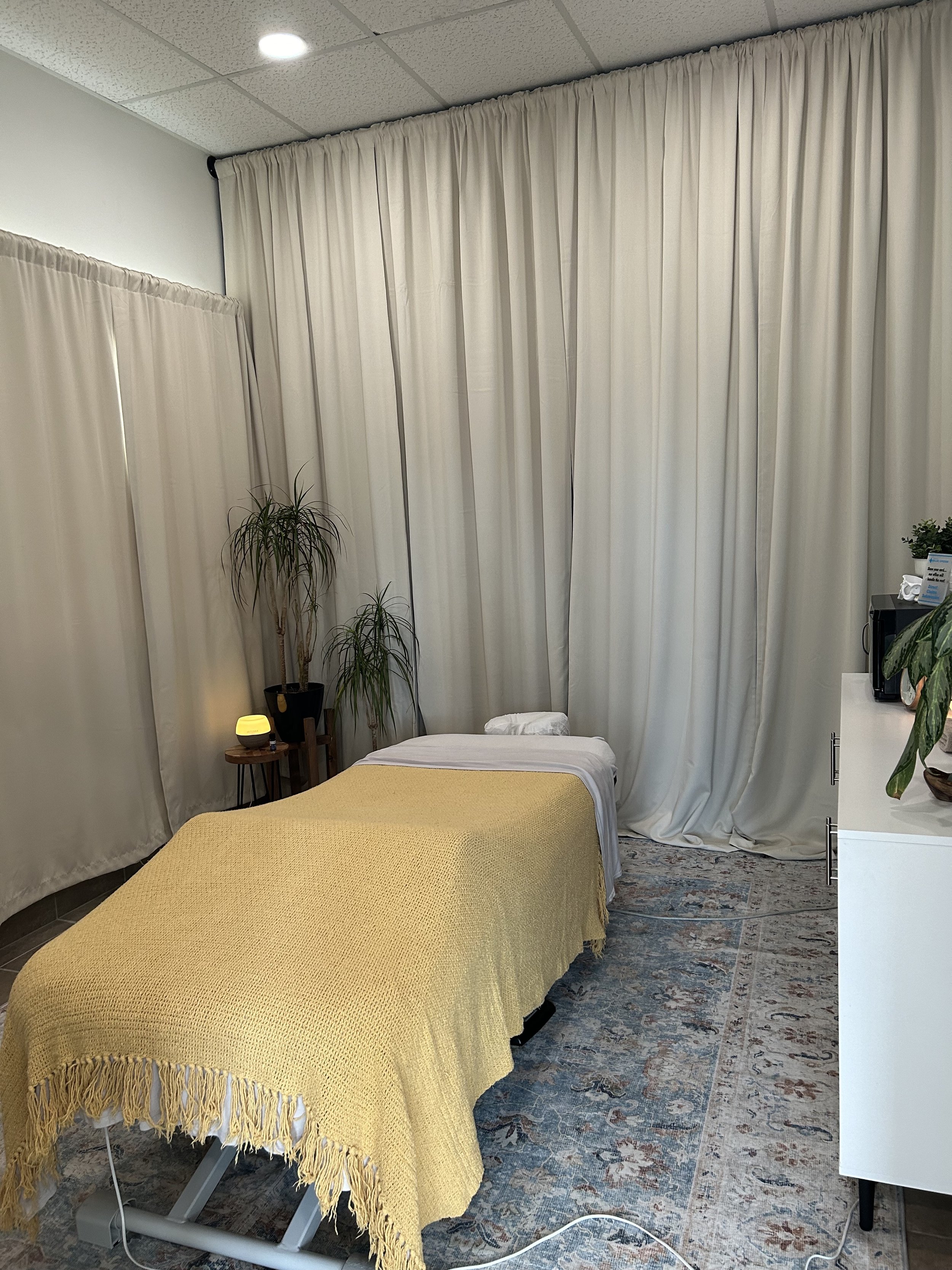 Stony Plain Massage. Spruce Grove Massage. Massage therapy room with a massage table covered by a yellow blanket, surrounded by curtains and indoor plants, soft lighting, and a patterned rug.