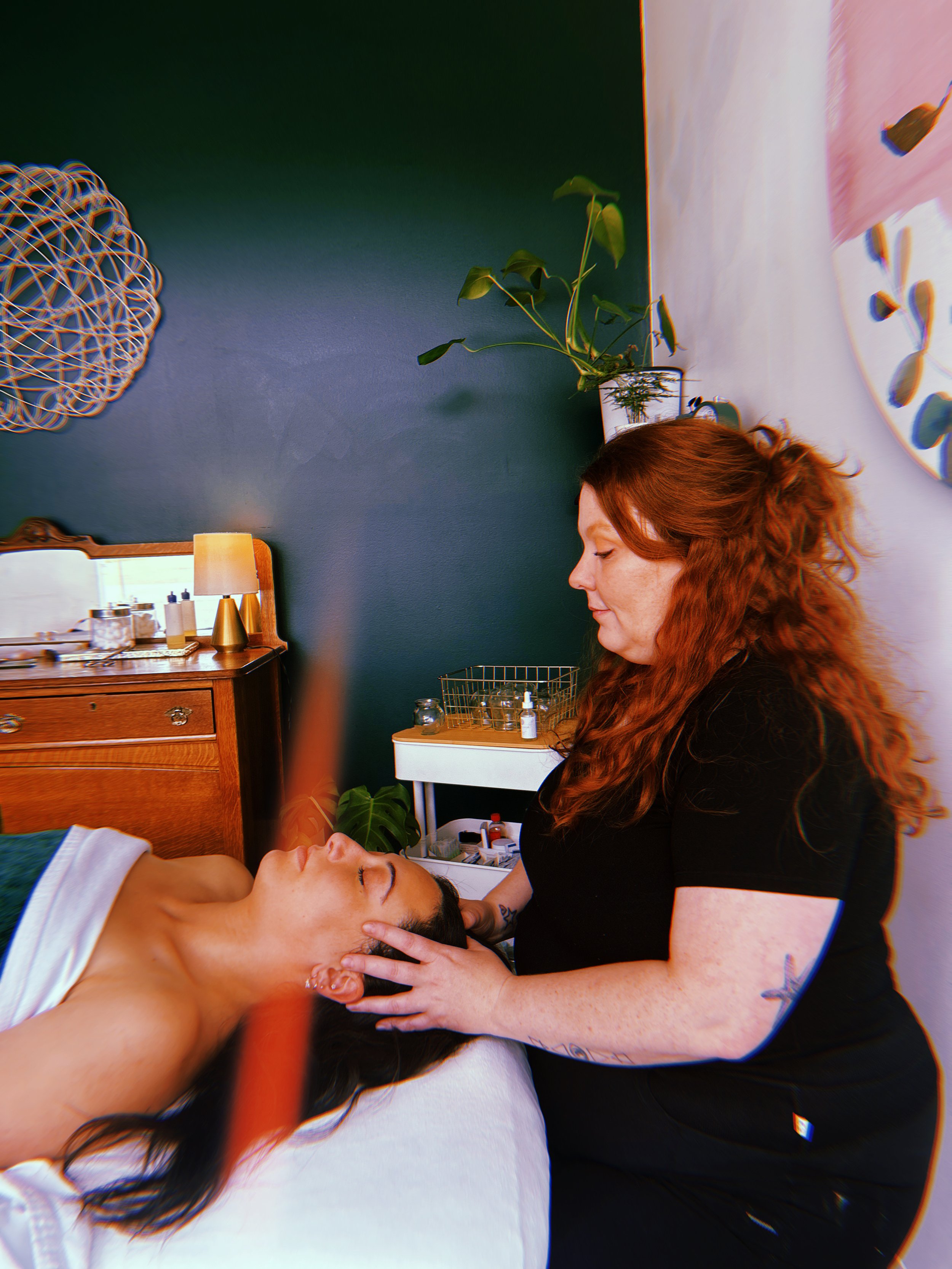 The Garden Holistic. YEG. A massage therapist giving a head massage to a client lying on a treatment bed in a dimly lit room with green walls and plants.