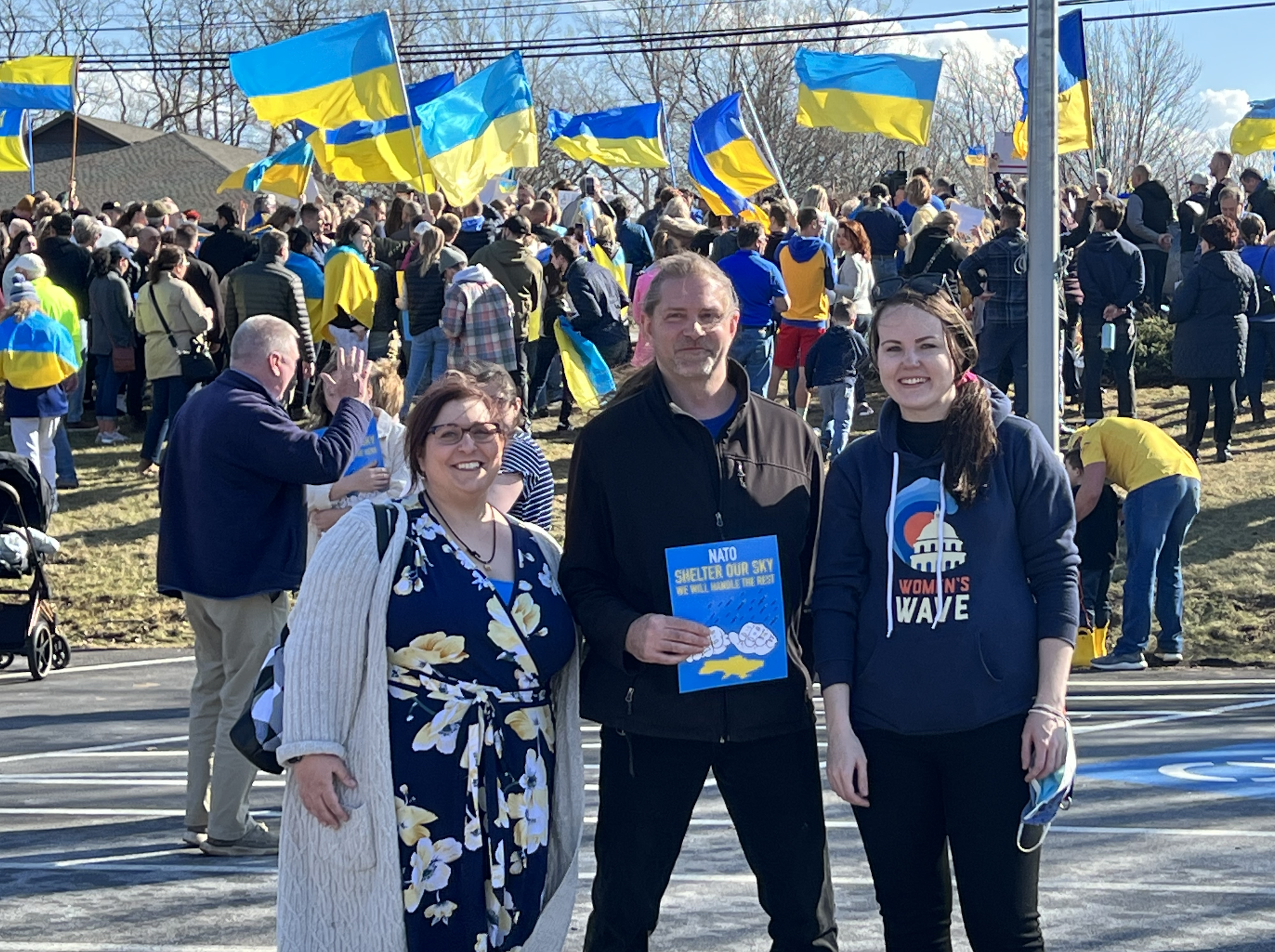 A diverse group of people at an outdoor rally holding and waving Ukrainian flags. Three individuals in the foreground smiling at the camera, one holding a sign about NATO. Many attendees in the background, also holding flags, with a sunny sky overhead.
