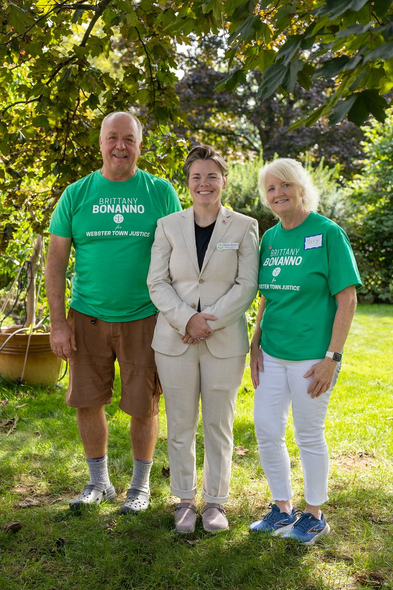 Three people standing outdoors on a grassy lawn under a leafy tree. The person in the middle is wearing a beige suit and glasses, and the two people on either side are wearing green T-shirts with political campaign messages. The woman on the right has a name tag that says 'Mary' and is dressed in white pants and sneakers. The man on the left is in brown shorts and crocs. All are smiling at the camera.