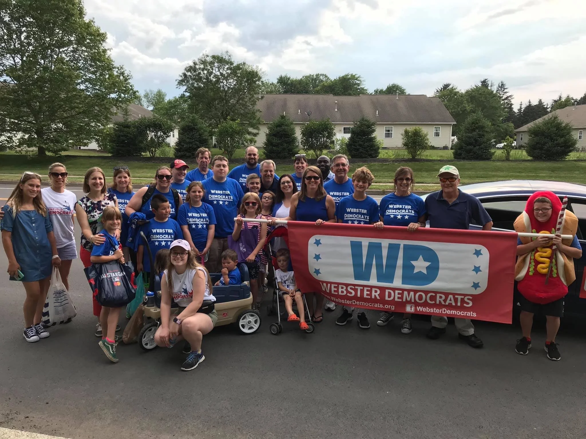 Group of people holding a banner that reads 'Webster Democrats' on a street with houses and trees in the background. The group includes adults and children, some wearing blue t-shirts with political slogans, and one person dressed as a hot dog hotdog mascot.